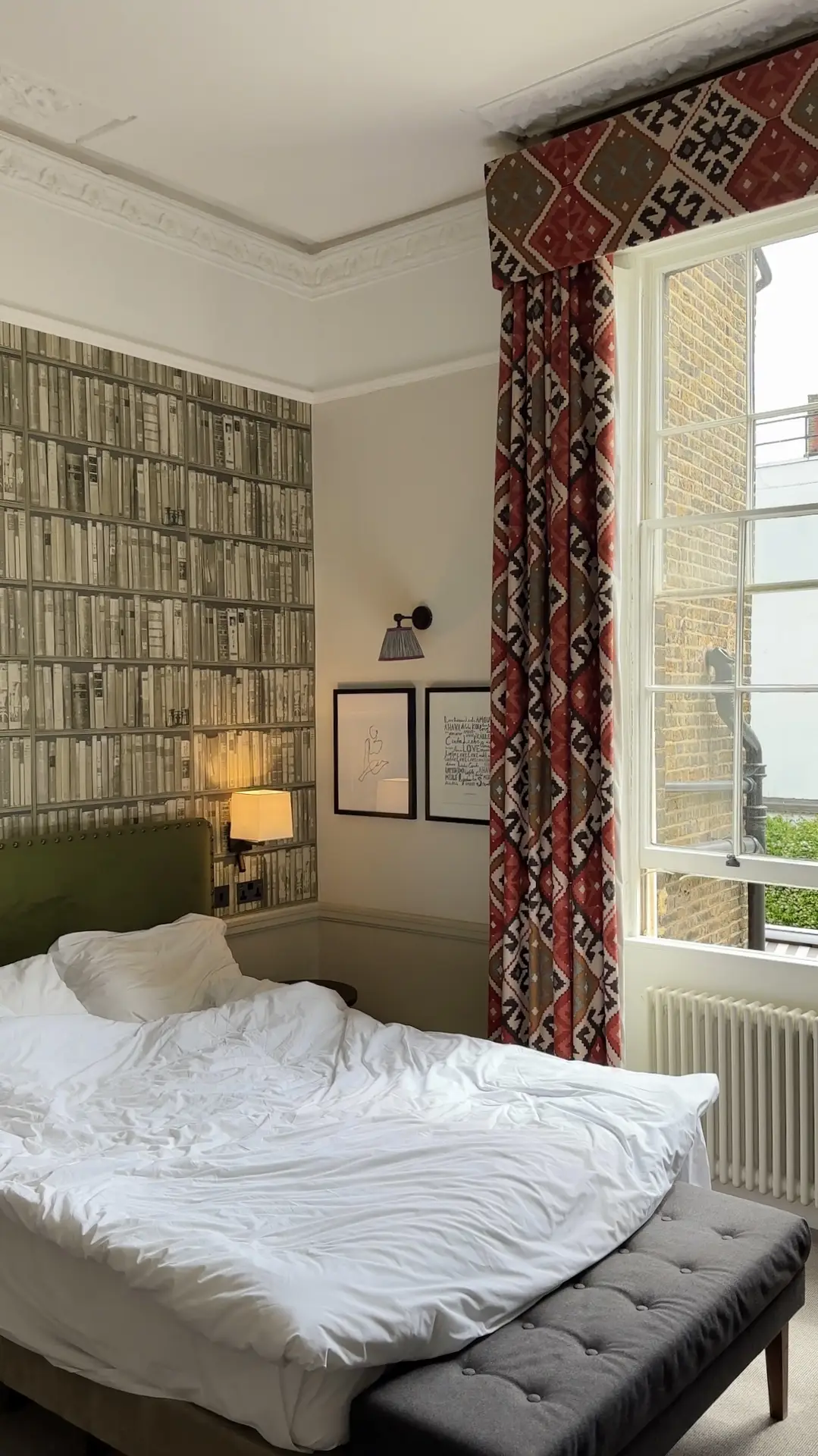 Eclectic bedroom featuring a warm neutral paint color for the bedroom paired with a striking patterned wallpaper that mimics stacked books. Bold red and brown geometric curtains frame the window, adding contrast to the otherwise soft tones of the space. The simple bed with white bedding, green headboard, and modern wall lights creates a cozy yet character-filled atmosphere.