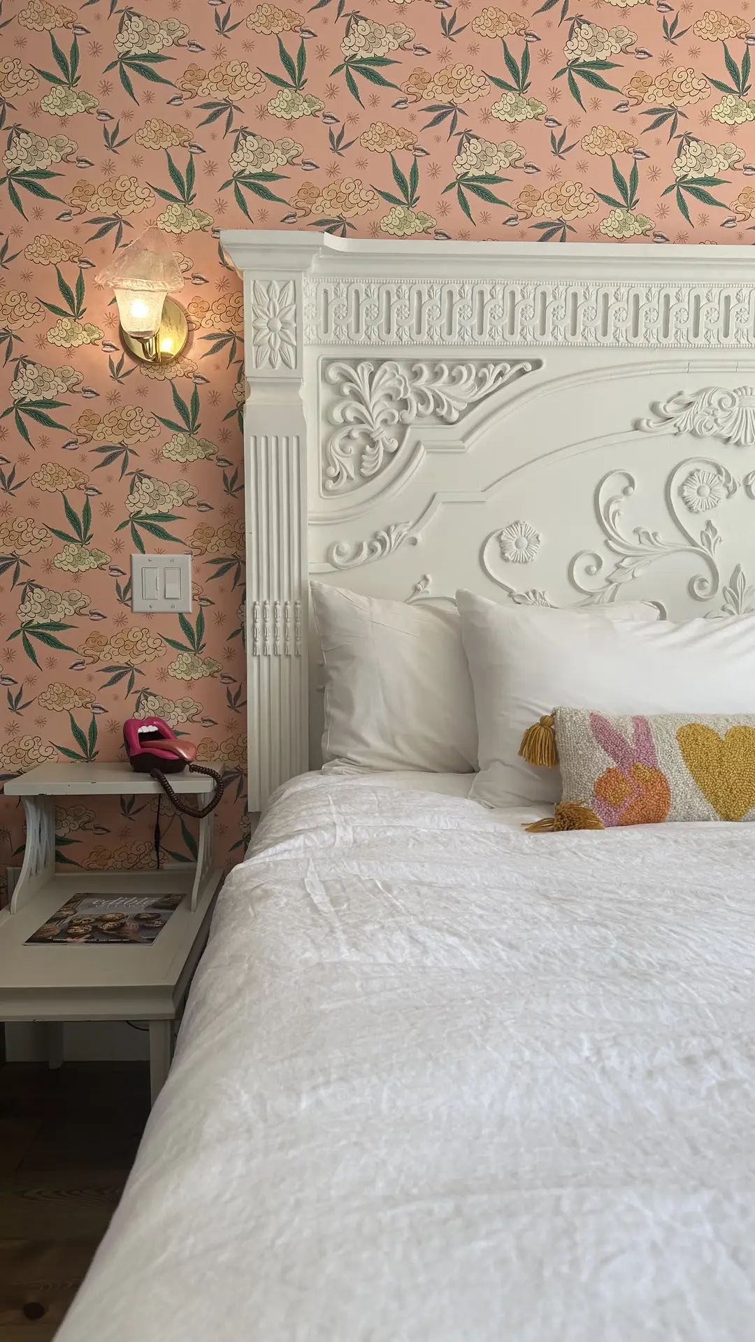 Playful bedroom featuring a bold paint color for the bedroom with pink floral pattern wallpaper, adding charm and personality to the space. A white ornate headboard with intricate detailing contrasts beautifully with the lively wall design. Crisp white bedding and a decorative cushion with textured heart and peace sign motifs complete the eclectic yet cozy look.