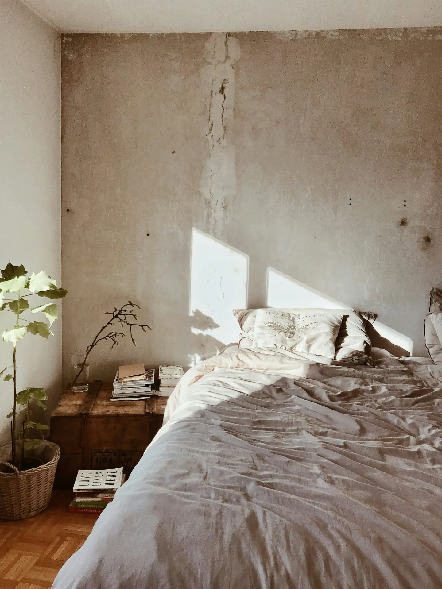 Minimalist bedroom showcasing a paint color for the bedroom in muted neutrals, paired with a raw wall for industrial charme that adds character and texture. Soft bedding in earthy tones complements the rugged backdrop, while natural light enhances the cozy yet edgy vibe. A wooden nightstand with stacked books and a potted plant bring warmth and balance to the space.