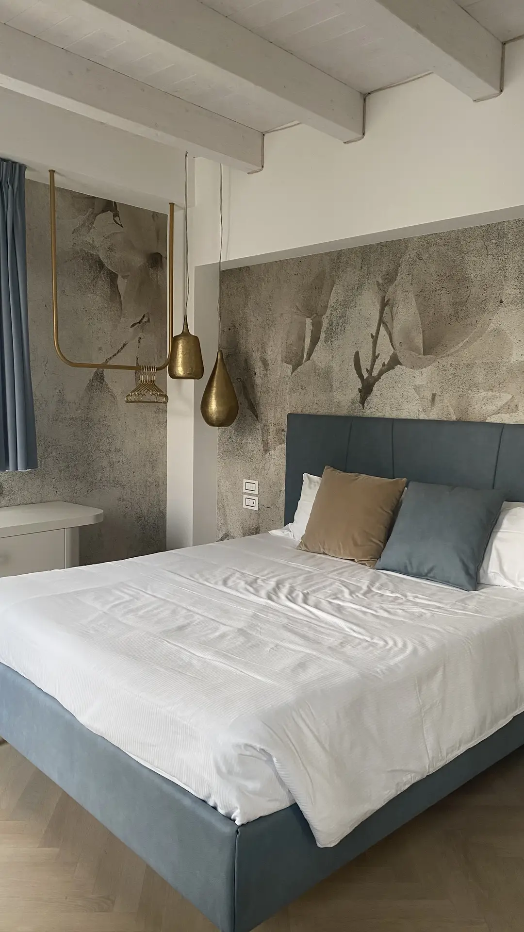 Contemporary bedroom highlighting a unique paint color for the bedroom with a textured trowel technique on the feature wall, giving the space depth and artistic flair. The room includes a blue upholstered bed with neutral-toned pillows, paired with gold pendant lights that add a touch of elegance. Soft blue curtains and wooden ceiling beams balance modern design with a cozy atmosphere.