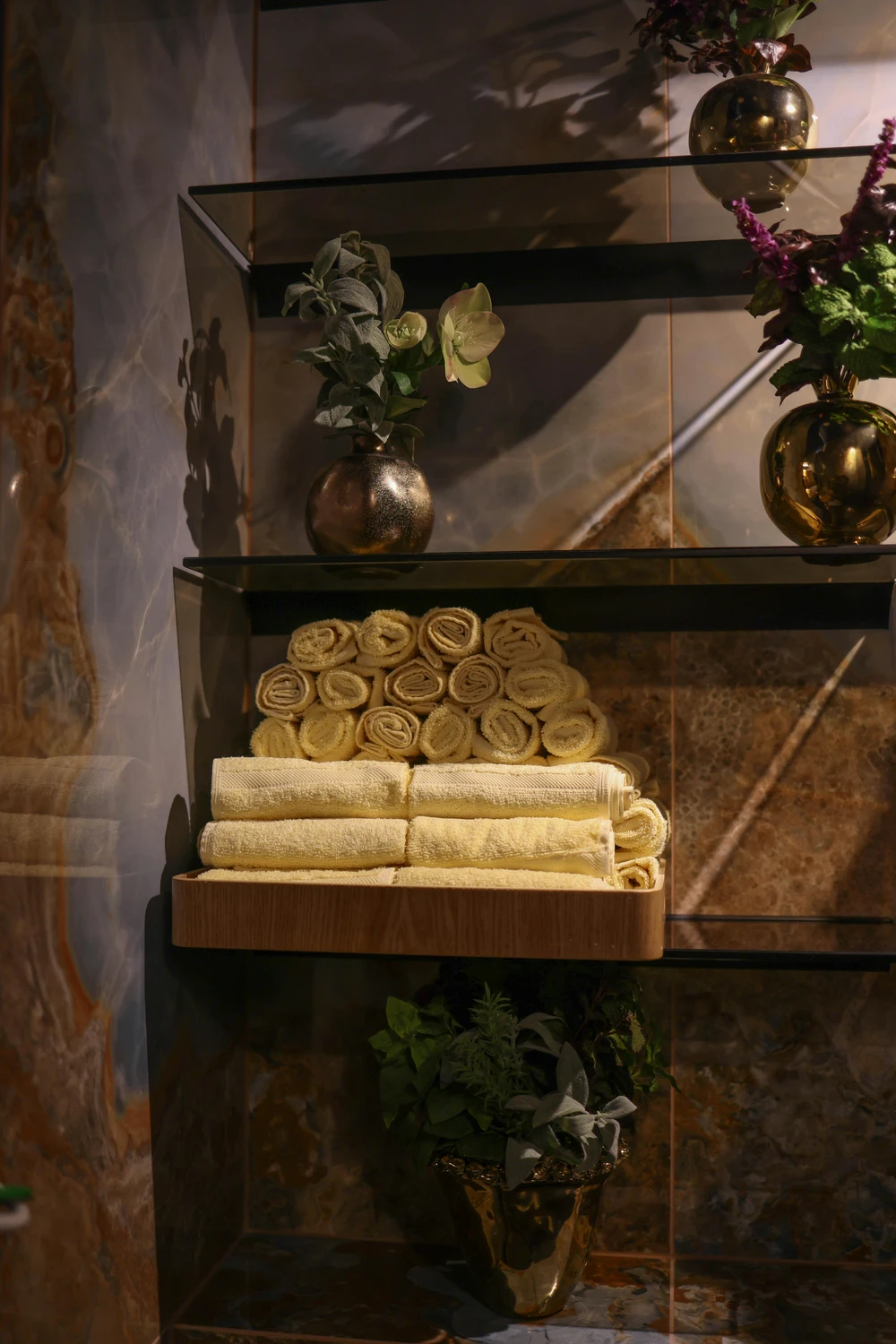 Elegant bathroom corner with sleek floating shelves, styled with rolled yellow towels and decorative gold vases filled with greenery and flowers, offering stylish small bathroom ideas. The warm marble wall backdrop enhances the cozy spa-like atmosphere while maximizing storage in a compact space.
