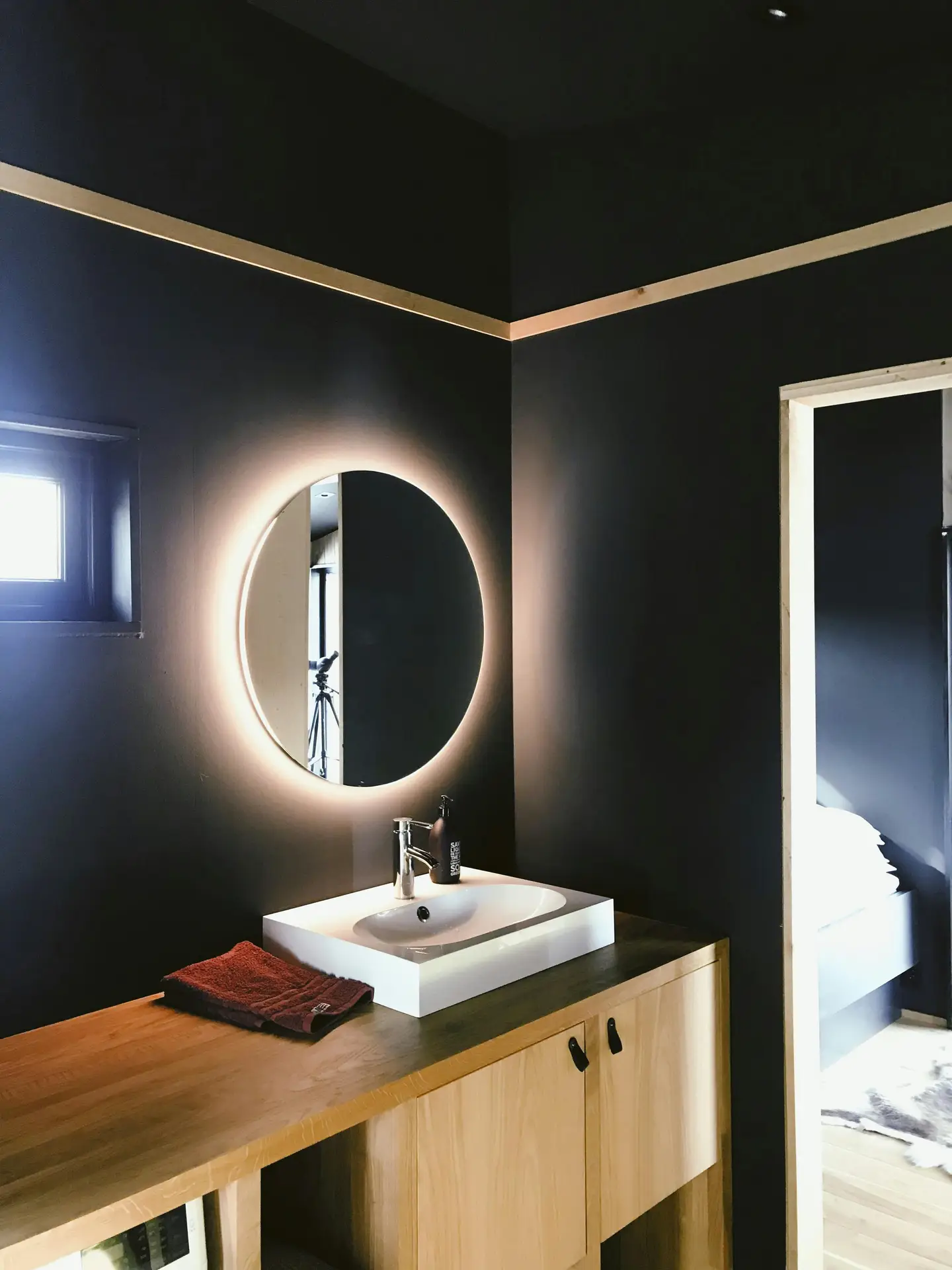 A sleek vanity with wood cabinetry, a white vessel sink, and a round backlit mirror on dark-painted walls, showing how clever lighting can elevate a small bathroom remodel with both style and function.