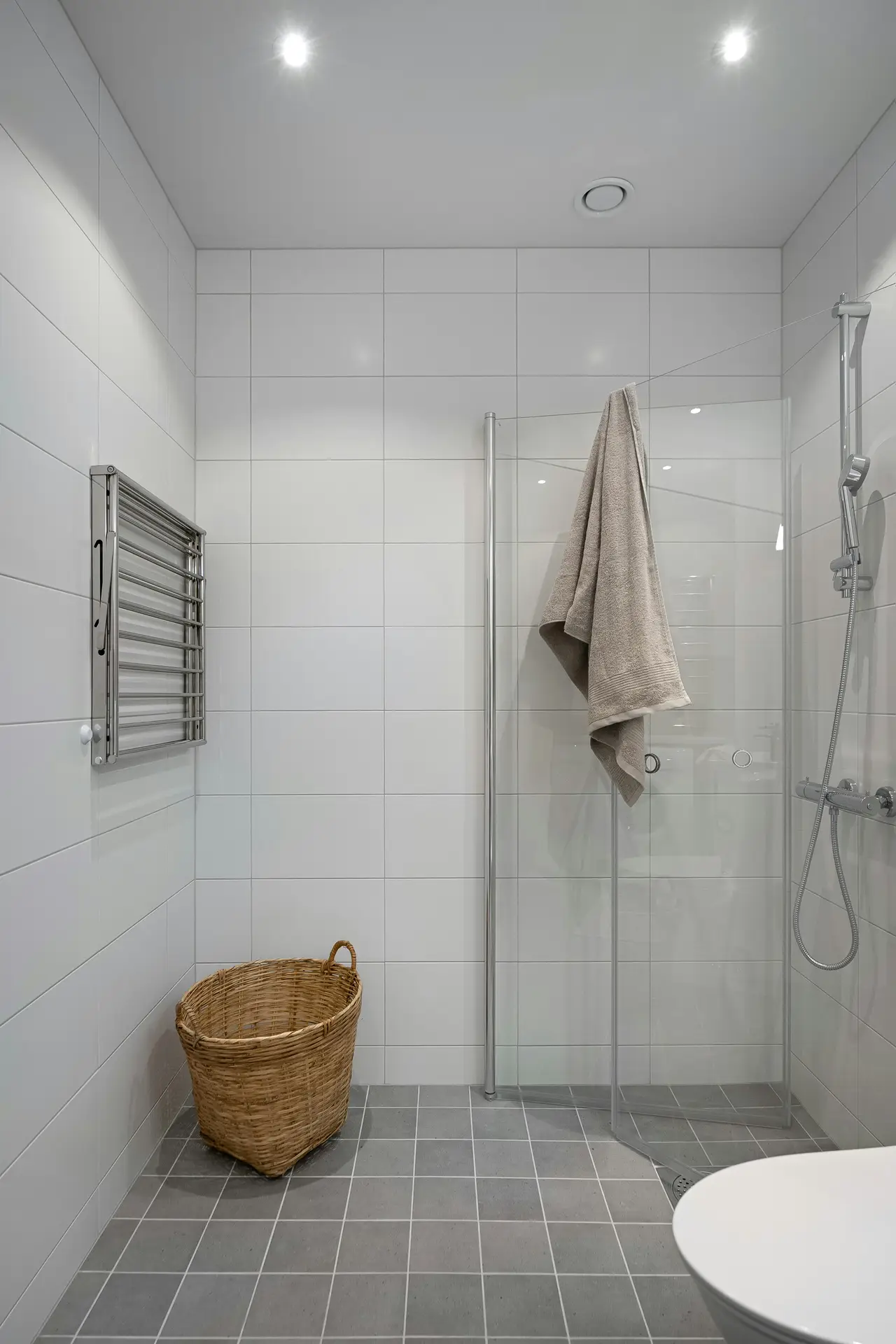 Modern small bathroom remodel with white tiled walls, gray floor tiles, and a sleek glass corner shower featuring a rainfall and handheld fixture. A wicker laundry basket and heated towel rack complete the design, making the space functional while maintaining a clean, minimalist look.