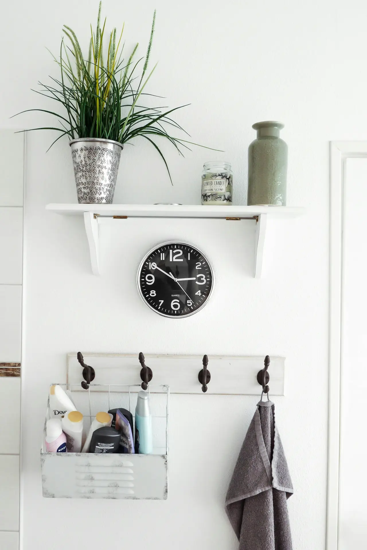 Wall decor in a small bathroom remodel featuring a simple hook system with dark metal hooks for towels and storage baskets, a black wall clock, and a floating white shelf styled with plants, a candle, and a ceramic vase for a clean yet functional look.
