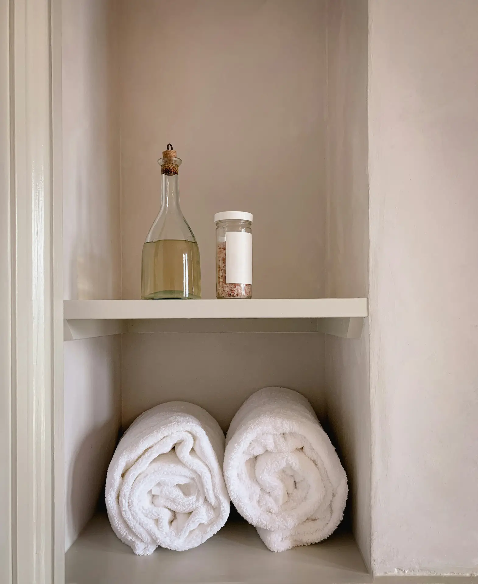 Built-in shelving with rolled white towels, bath salts, and a glass bottle, demonstrating how to incorporate smart storage solutions in a small bathroom remodel while keeping the look minimal and spa-inspired.