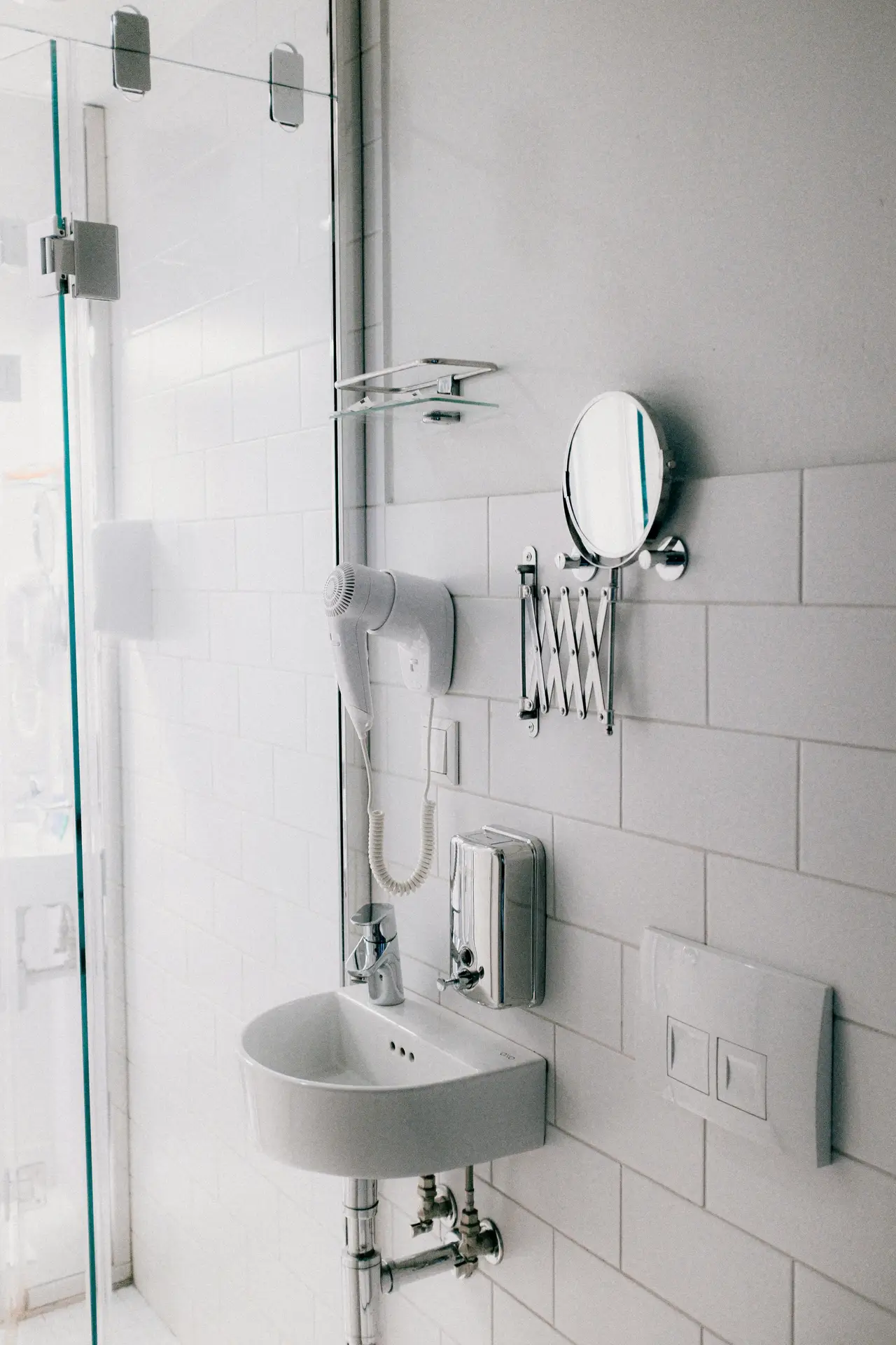 Compact small bathroom remodel featuring a wall-mounted sink, glass shower enclosure, and clever storage with mirror and hairdryer holders. The design uses tiles halfway up & damp-proof paint above, creating a clean, durable, and space-efficient solution perfect for modern minimalist spaces.