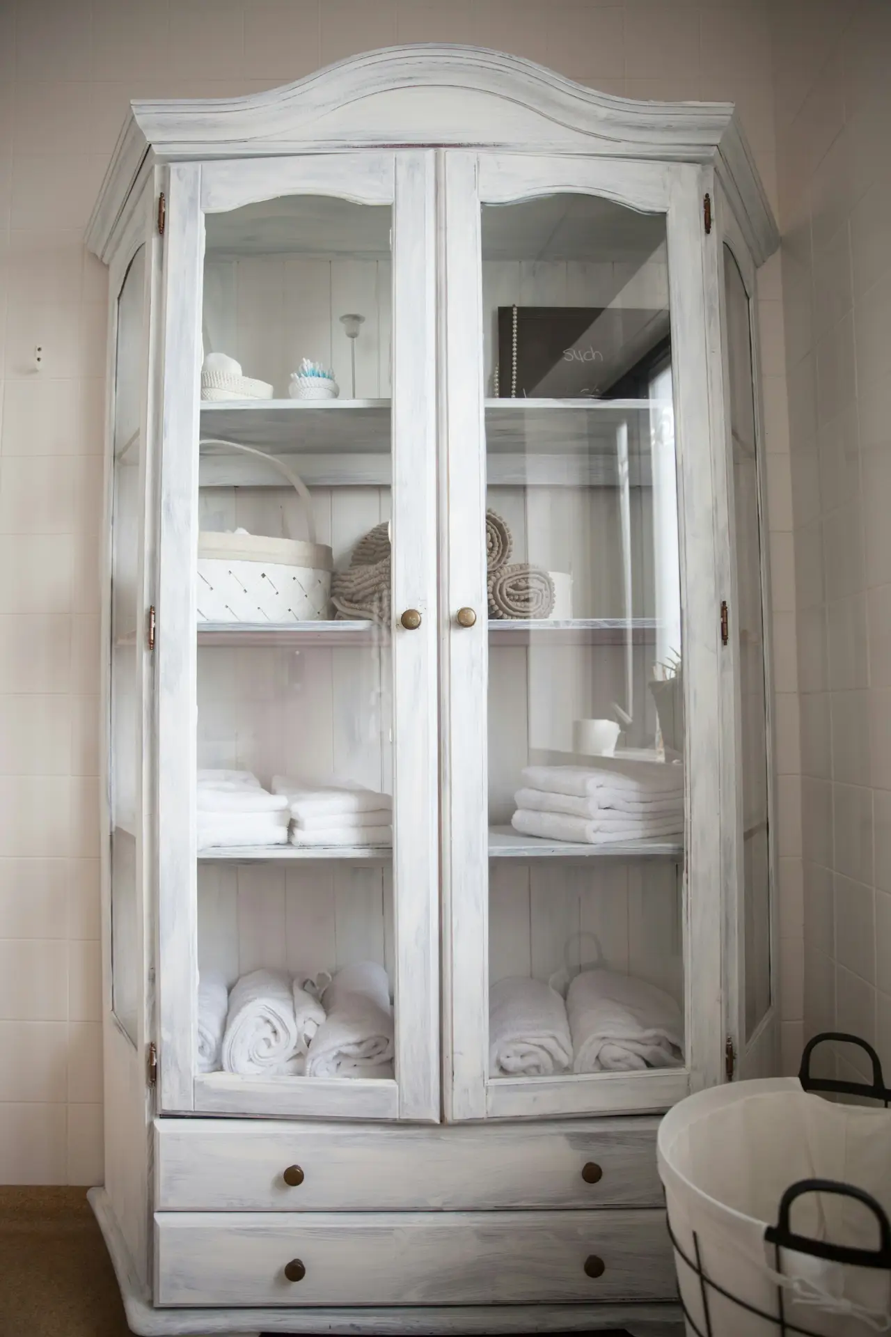 A shabby-chic glass-front vertical cabinet styled with neatly folded and rolled towels, baskets, and small bathroom essentials, illustrating clever storage for a small bathroom remodel while keeping the design light and airy.