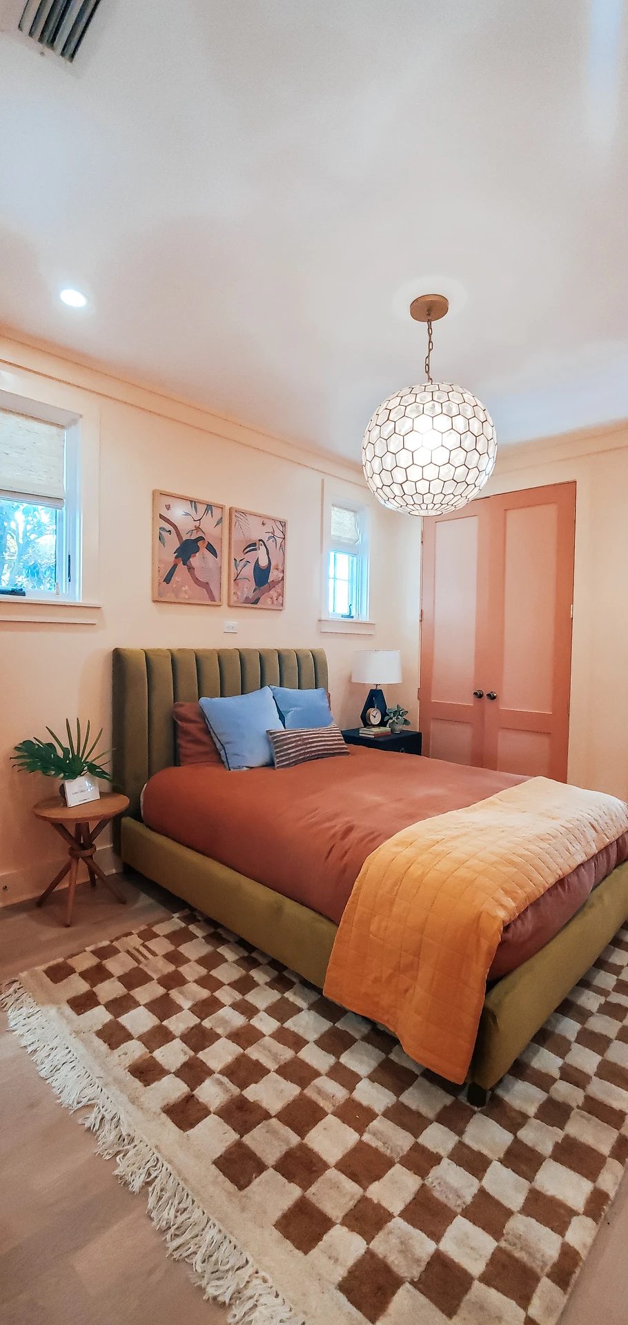 A playful take on small bedroom decor where warm tones dominate the space, featuring an olive-green upholstered bed with layered pillows in blue and brown. The checkered rug is styled with the carpet 2/3 under the bed, adding visual depth and balance to the layout. Peach-toned walls, matching closet doors, and a honeycomb pendant light bring harmony while framed bird artwork and plants on side tables add a cozy, personal touch.