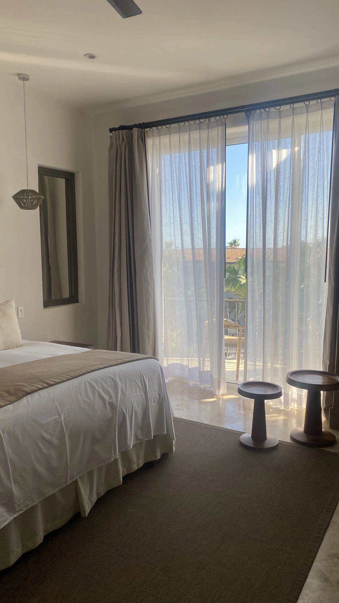 Elegant small bedroom decor with a neatly made bed in neutral tones, complemented by soft floor to ceiling curtains that frame a large glass door opening to a balcony. Natural light floods the room, while minimalist wooden side tables and a simple pendant lamp complete the airy, modern look.