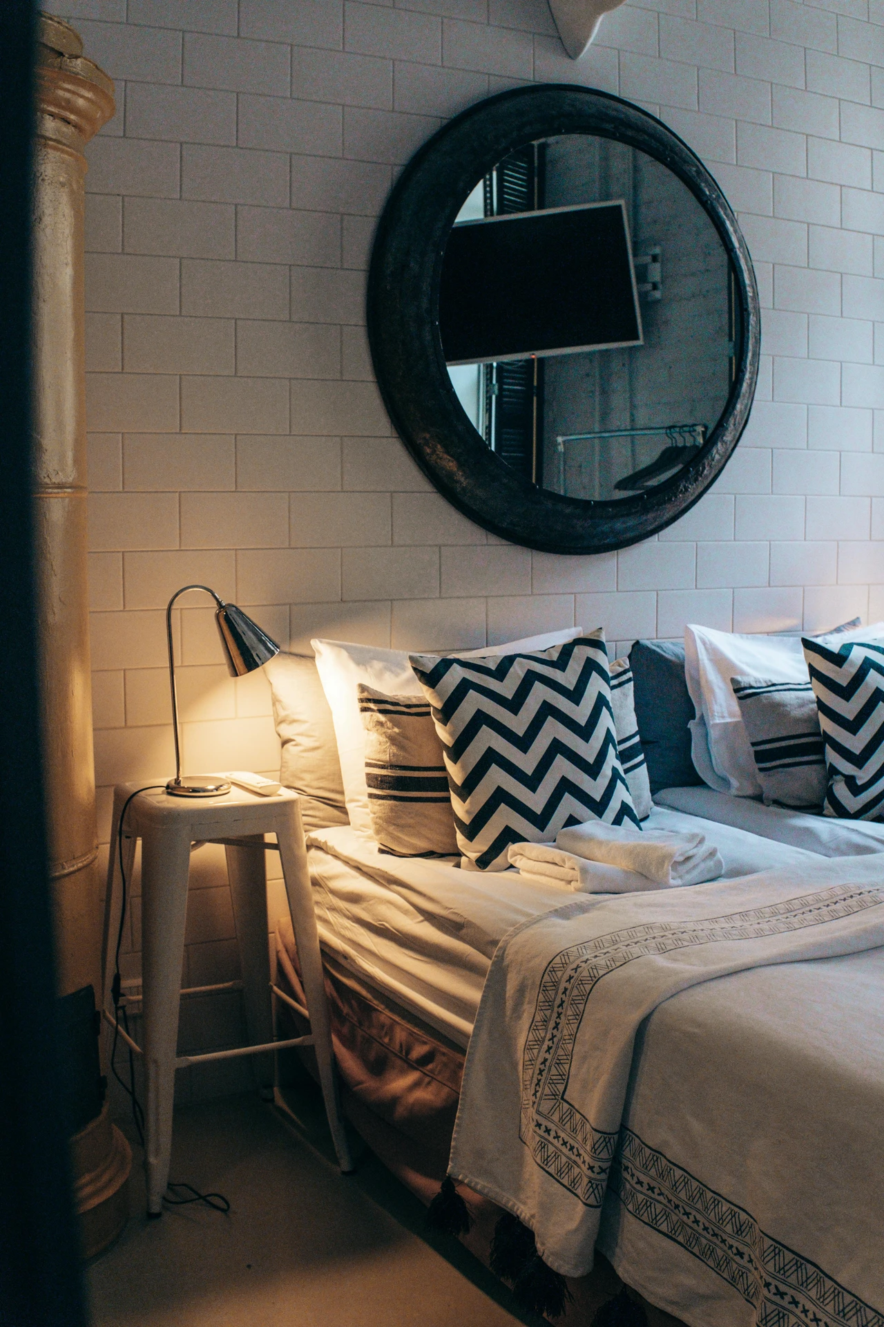 Cozy small bedroom decor with a low-profile bed dressed in white linens and patterned cushions in chevron and striped designs. A simple stool doubles as a nightstand with a sleek lamp, while a large round wall mirror above the bed adds depth and character to the tiled accent wall.