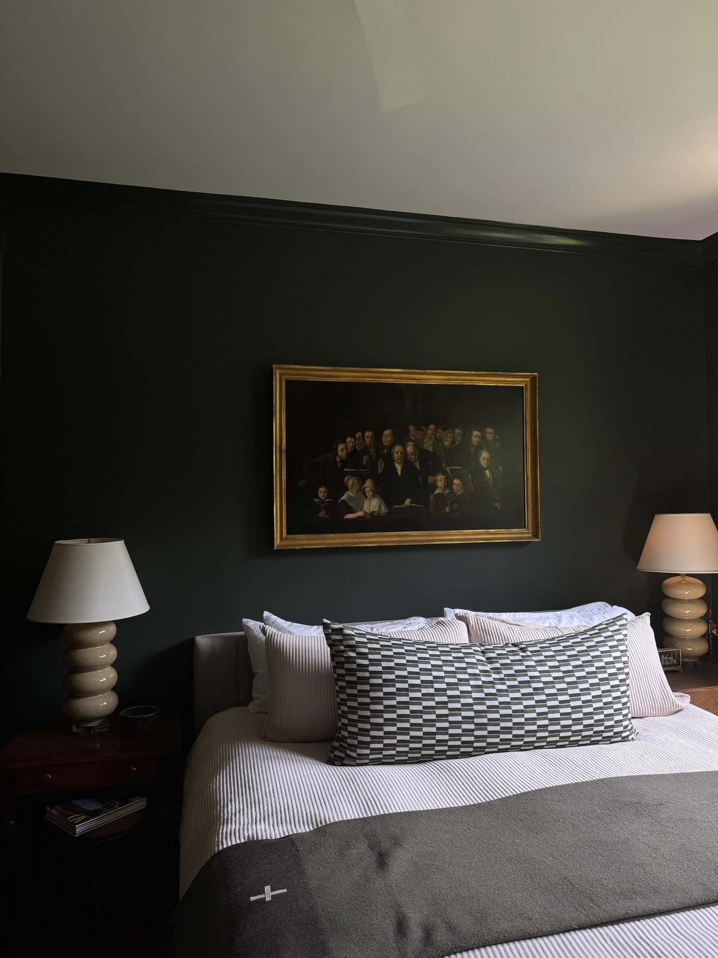 Elegant small bedroom decor featuring a dark painted accent wall with a classic framed painting as the focal point. The bed is layered with crisp white linens and highlighted by a bold statement lumbar pillow in a geometric black-and-white pattern, flanked by sculptural lamps on each nightstand. The combination of rich tones, symmetry, and texture adds depth and sophistication to the intimate space.