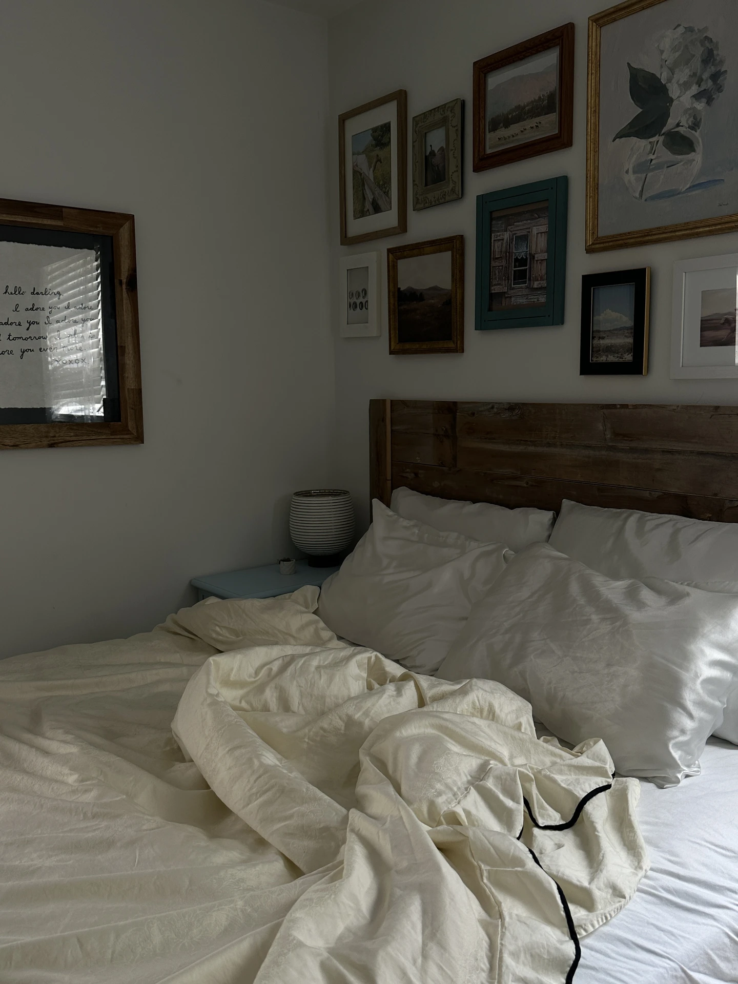 Cozy small bedroom decor with a rustic wooden headboard and soft cream bedding casually draped for a relaxed feel. The wall above the bed features a charming mini gallery above bed, mixing paintings, photos, and sketches in frames of different sizes and finishes. This layered arrangement creates warmth and personality in a compact sleeping space.