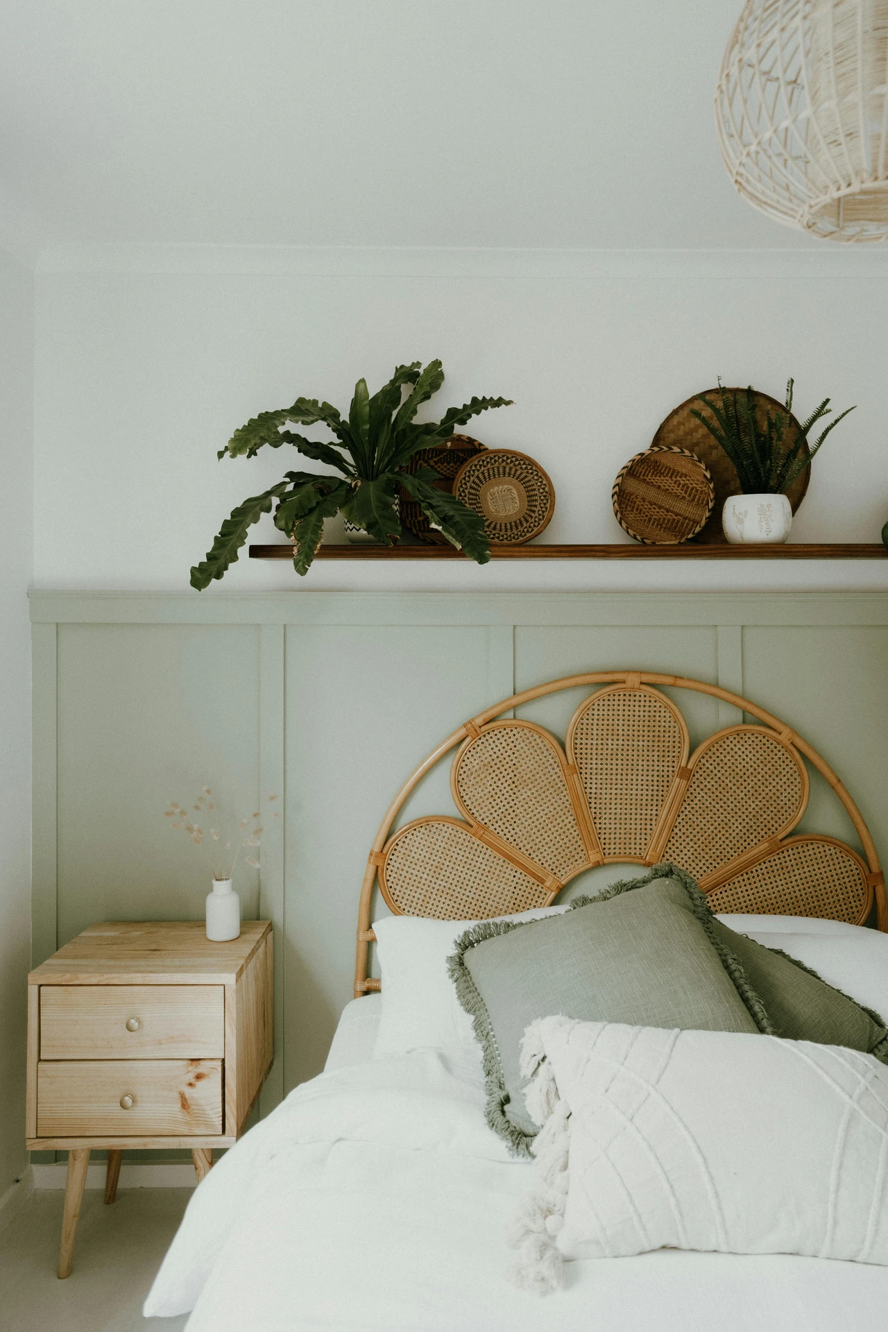 A light and airy small bedroom decor concept featuring a rattan headboard in a floral design, styled with soft white and sage pillows. Above the bed, woven baskets and potted plants add minimal greenery for a natural touch without overwhelming the space. A wooden nightstand with a simple white vase keeps the look warm, calm, and cohesive.