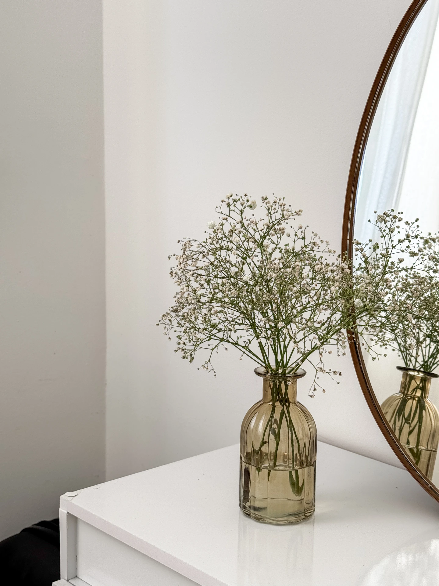 Minimalist small bedroom decor with a round mirror reflecting soft light and a simple white dresser topped by a delicate glass vase. The airy arrangement of baby's breath creates vertical accents with tall vases, bringing elegance and height into the compact space. The clean lines and subtle natural touch highlight how even small details can elevate a serene, modern bedroom.