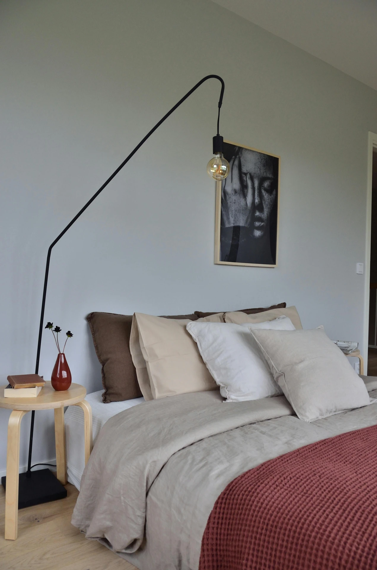 A serene example of small bedroom decor where soft neutrals dominate the space, creating a calming atmosphere. The bed is styled with tone-in-tone bedding and one texture layer, featuring beige and cream pillows, light taupe sheets, and a rust-colored knitted throw for depth. A minimalist setup with a slim bedside table, books, and a single red vase ties the look together under a bold black lamp and monochrome wall art.