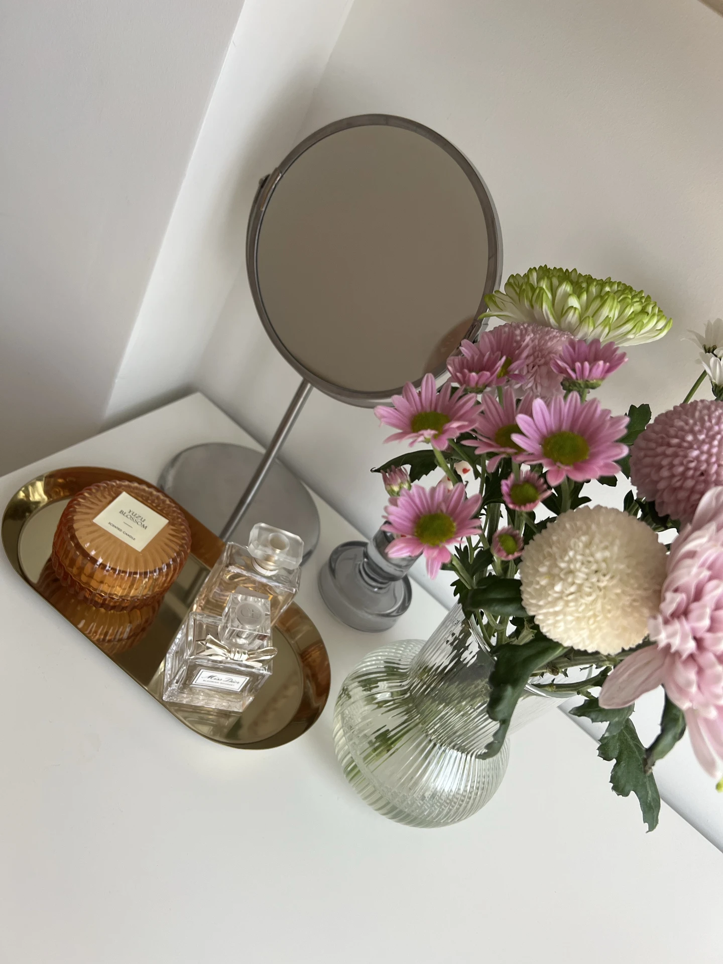A bright and fresh small bedroom decor vignette featuring a vanity corner with delicate tray styling. On the golden tray sit a scented candle and elegant perfume bottles, complemented by a glass vase filled with soft pink and white flowers. A simple tabletop mirror completes the cozy yet chic arrangement, adding both practicality and charm.