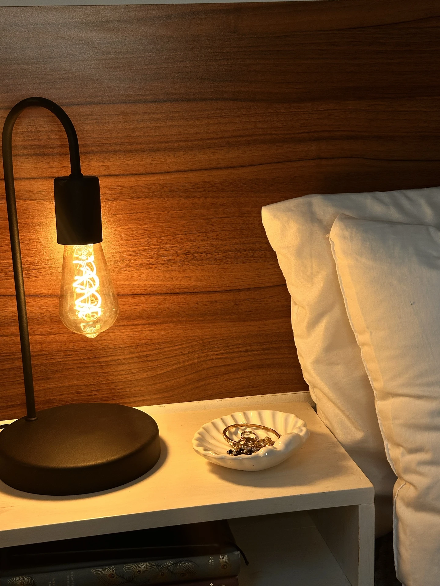 Warm small bedroom decor with a sleek black lamp casting a cozy glow beside crisp white pillows. A compact wall-mounted bedside table holds a shell-shaped dish with jewelry, balancing function and style against the rich wooden headboard.