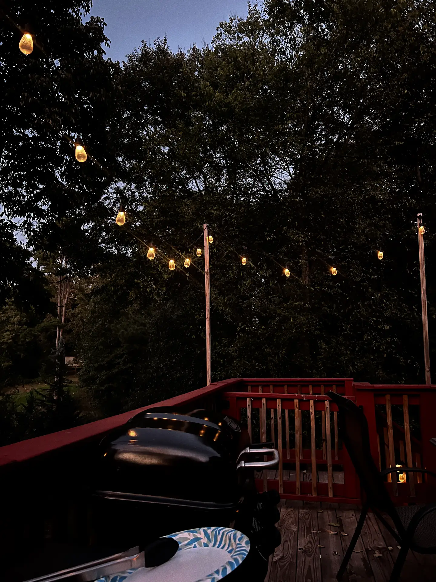 Cozy evening patio scene featuring glowing fairy lights strung above a wooden deck surrounded by trees. A grill and outdoor chairs create a relaxed atmosphere, perfect for late summer dinners or small gatherings. A warm example of outdoor lighting ideas that add charm and ambiance to any backyard space.