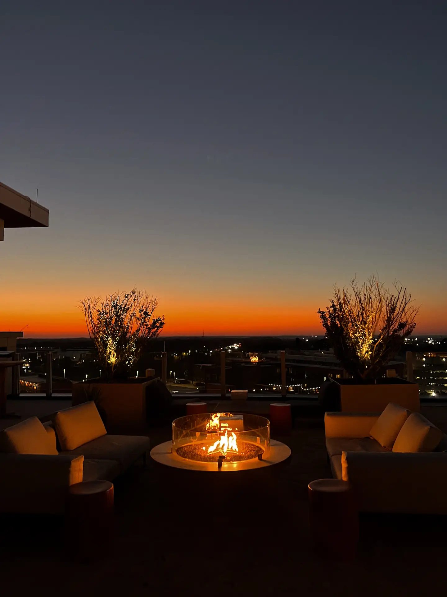 A modern rooftop terrace at sunset featuring a round glass-enclosed fireplace surrounded by comfortable lounge seating. The warm firelight contrasts beautifully with the fading orange sky and softly lit trees, showcasing elegant outdoor lighting ideas for cozy, stylish evening gatherings.