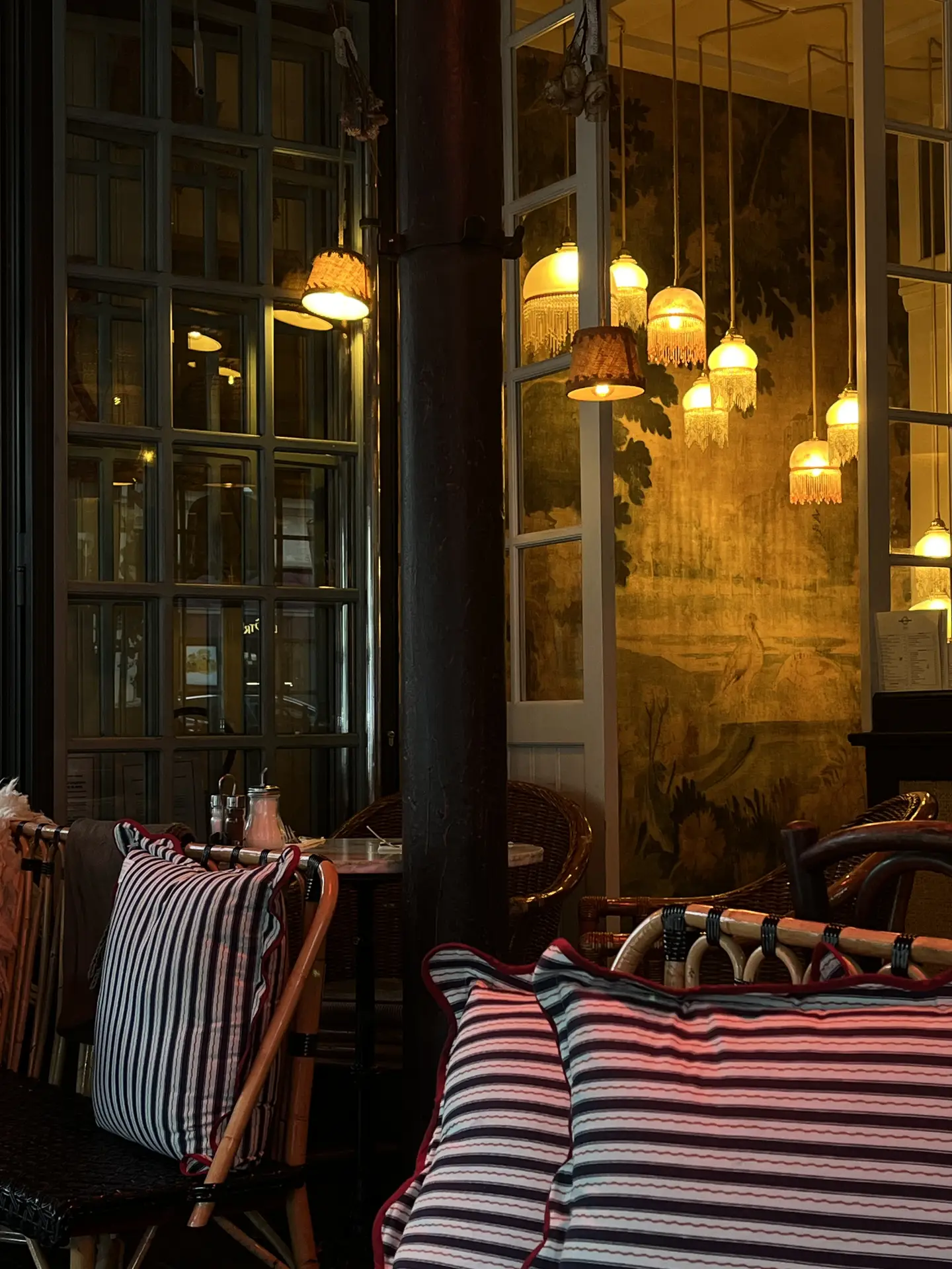 A cozy café corner with striped cushions, rattan chairs, and warm lighting that creates an inviting atmosphere. Several fringed fabric pendant lamps hang from the ceiling, casting a soft glow over the seating area. The scene captures charming outdoor lighting ideas for patios, bistros, or covered terraces with a vintage touch.