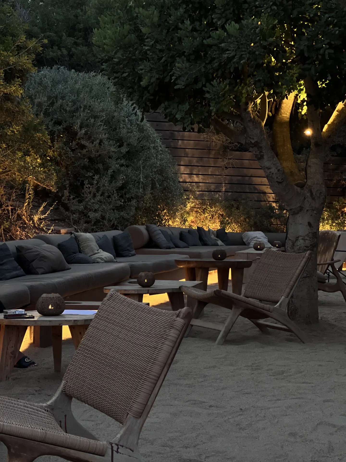 A stylish outdoor lounge with wooden chairs, cushioned benches, and a sandy ground, softly illuminated by indirect tree lights. The warm glow highlights the greenery and seating area, creating a cozy and inviting ambiance. A great example of outdoor lighting ideas that blend comfort and natural elements for relaxed evenings outside.