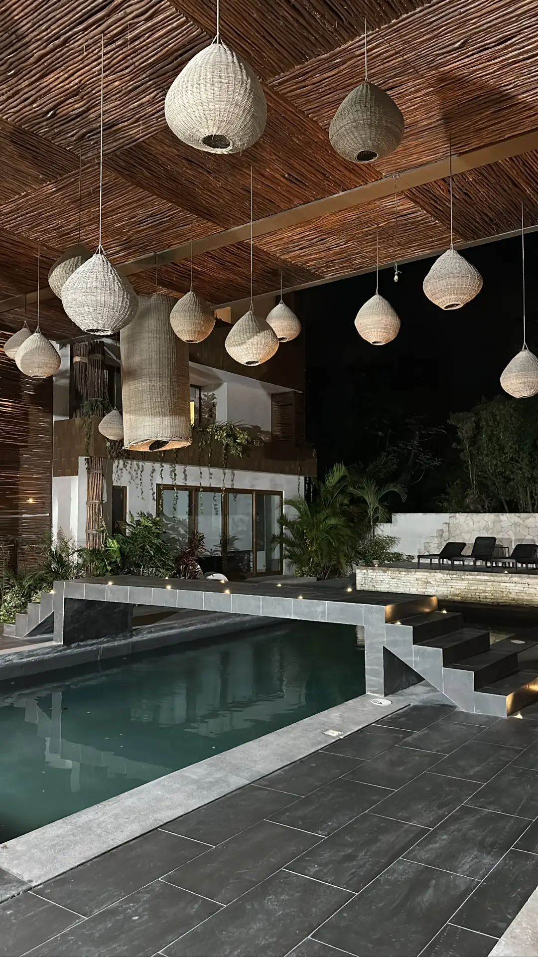 Stylish poolside area illuminated by warm lampions hanging from a rustic wooden ceiling. The space features sleek stone tiles, subtle stair lights, and lush greenery, creating a cozy yet elegant atmosphere. A perfect example of outdoor lighting ideas that blend natural textures with modern design for a serene evening ambiance.