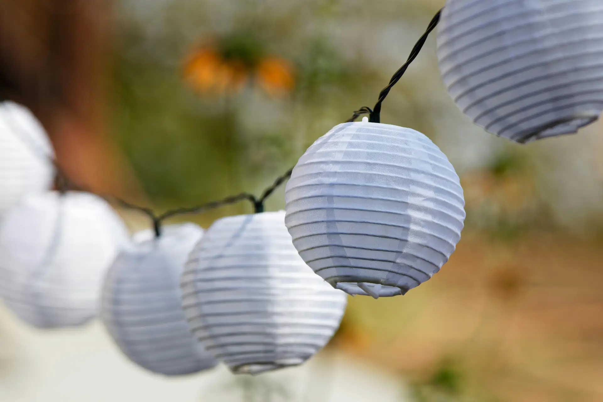 A close-up of white paper lanterns hanging on a string outdoors, softly illuminated in natural light. The background is slightly blurred, showing greenery and warm tones. This setup offers simple yet stylish outdoor lighting ideas that add a festive and cozy touch to gardens, patios, or evening gatherings.