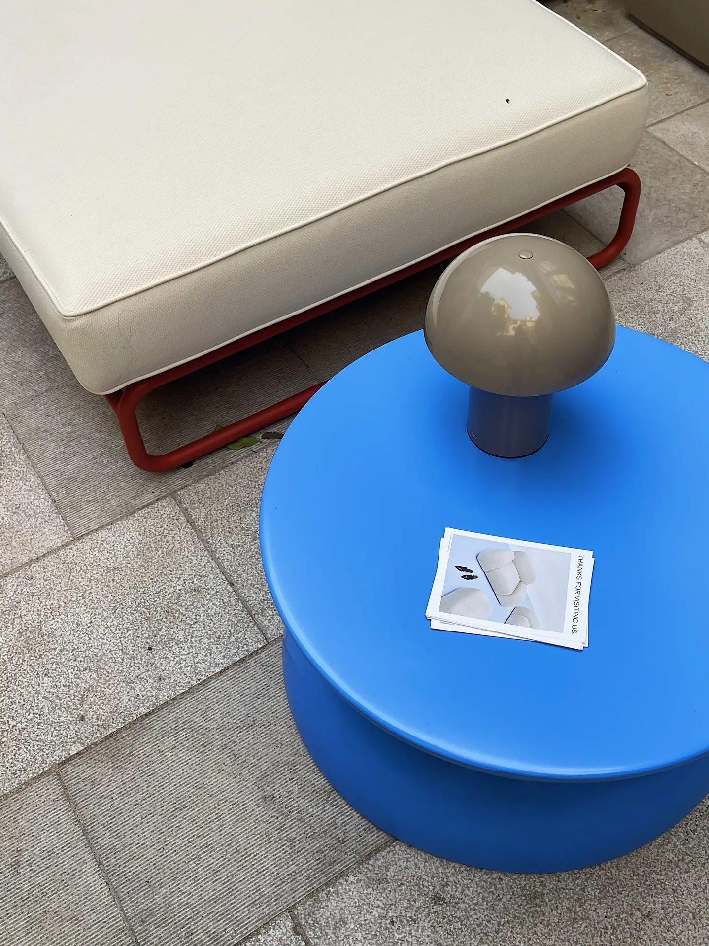 A modern outdoor setup with a bright blue round table holding a mushroom table lamp and a small stack of cards. Next to it sits a beige cushioned lounger with a red metal frame. The scene blends playful design with functionality, offering creative outdoor lighting ideas for contemporary patio or terrace spaces.