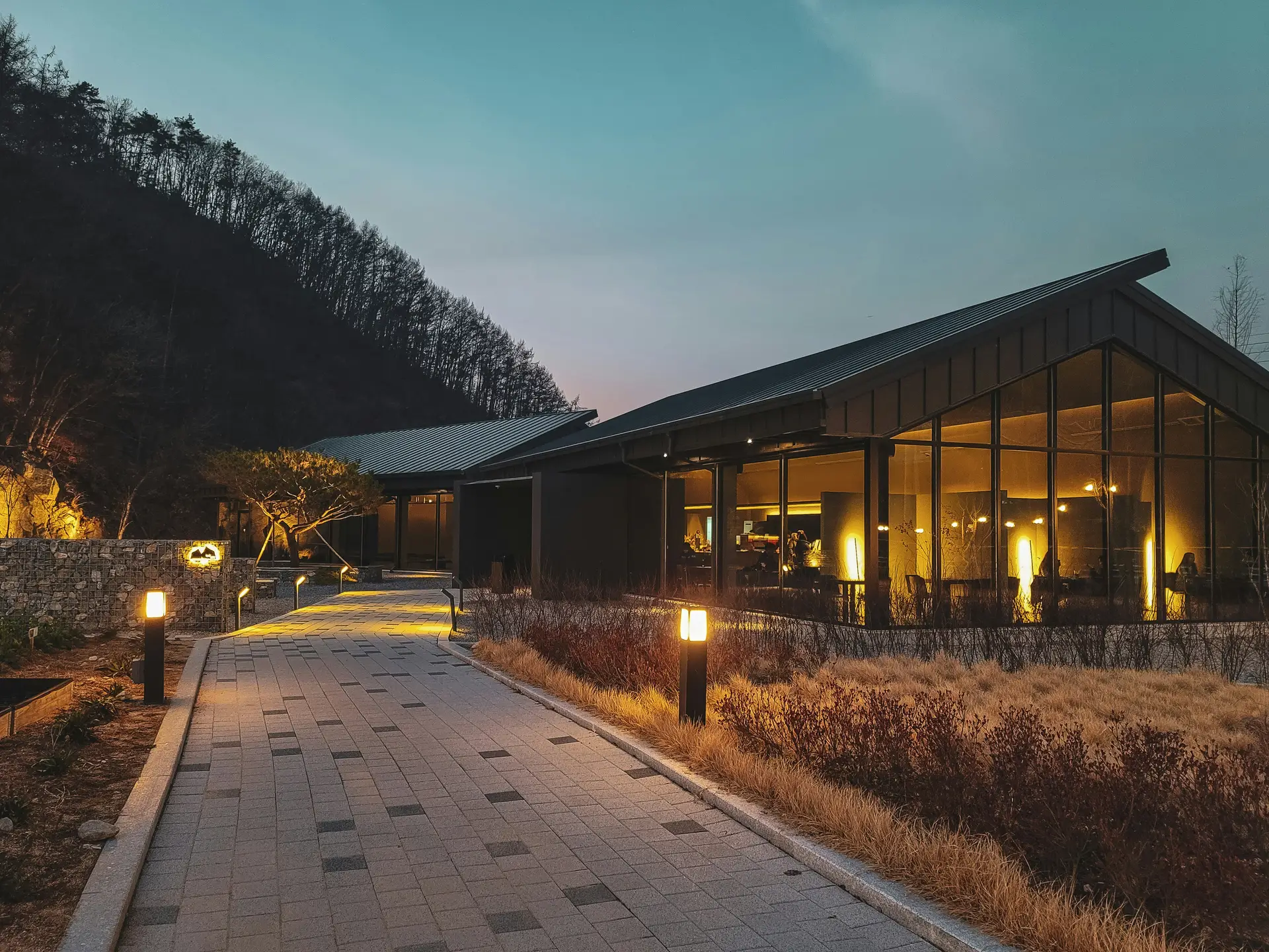 A modern building with large glass windows glows warmly at dusk, framed by surrounding hills and dry landscape plants. The walkway leading up to it is lined with sleek pathway bollard lights that softly illuminate the path. This scene illustrates elegant outdoor lighting ideas for safe and stylish walkways in contemporary settings.