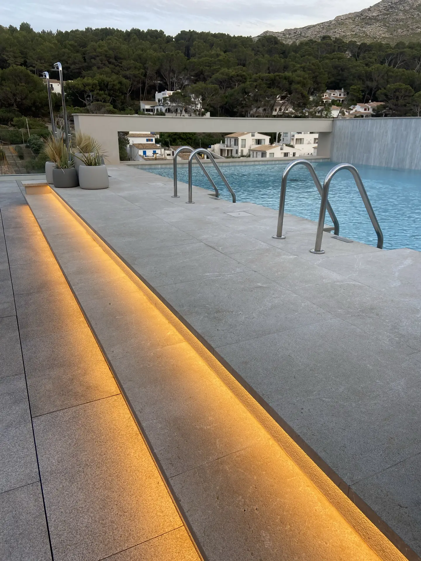 Modern poolside area with sleek stone flooring and elegant recessed step lights casting a soft golden glow along the pathway. Surrounded by lush greenery and minimalist architecture, this setting showcases stylish outdoor lighting ideas that enhance both safety and ambiance in contemporary spaces.