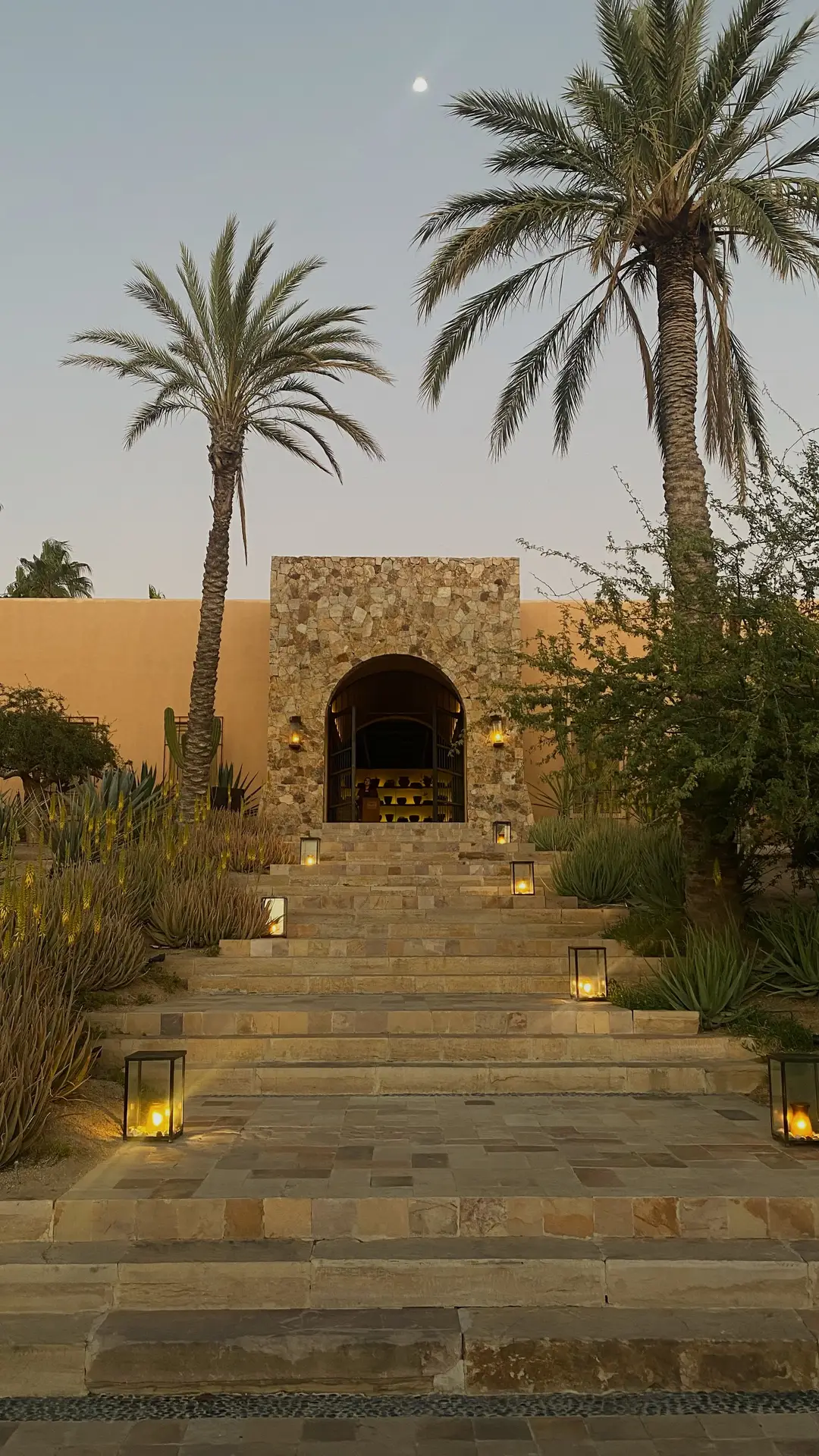A stone pathway lined with standing lanterns leads up to a rustic building framed by tall palm trees and desert plants. The warm glow of the lanterns enhances the earthy tones of the architecture, offering elegant outdoor lighting ideas for entrances or garden pathways in warm climates.