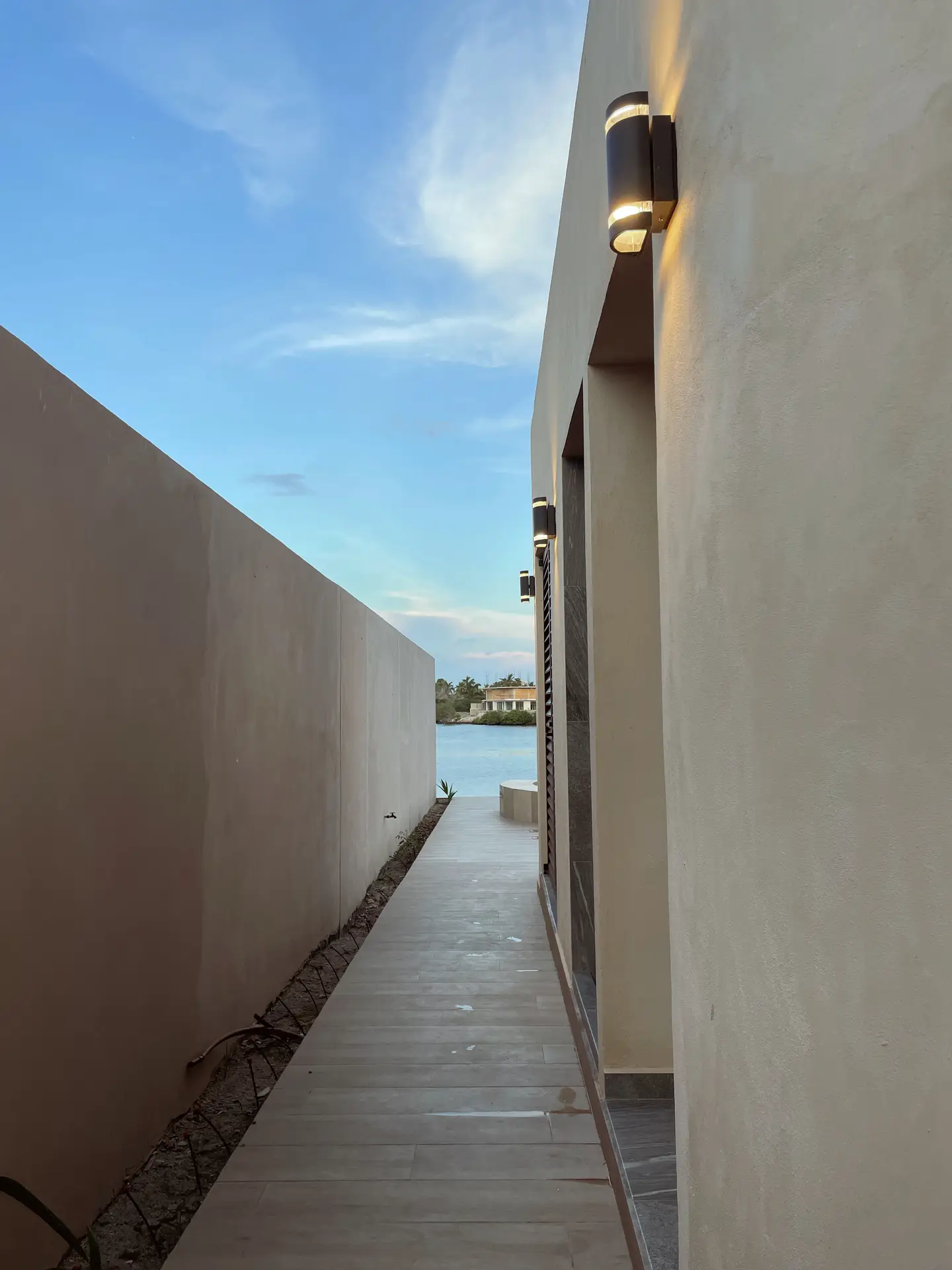 A narrow walkway between modern beige walls, leading toward a view of the water under a clear evening sky. Sleek up and down wall lights illuminate the passage with a soft glow, enhancing the minimalist architecture. This scene offers elegant outdoor lighting ideas for pathways or modern coastal homes.
