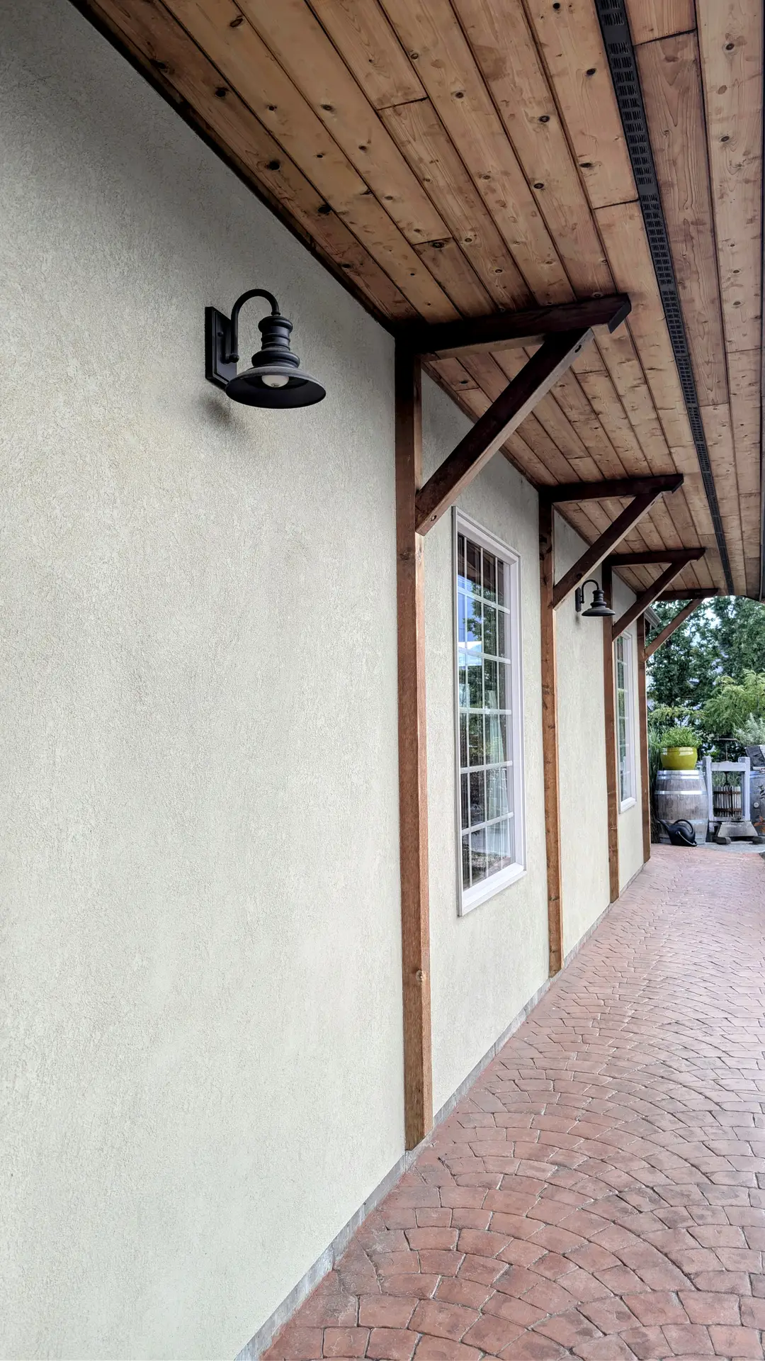 A covered walkway with wooden beams and a light-colored stucco wall featuring black metal wall sconces. The design combines rustic and modern elements, offering practical yet stylish outdoor lighting ideas for entryways, porches, or patios. The warm wood ceiling and brick pathway add a welcoming touch to the overall look.