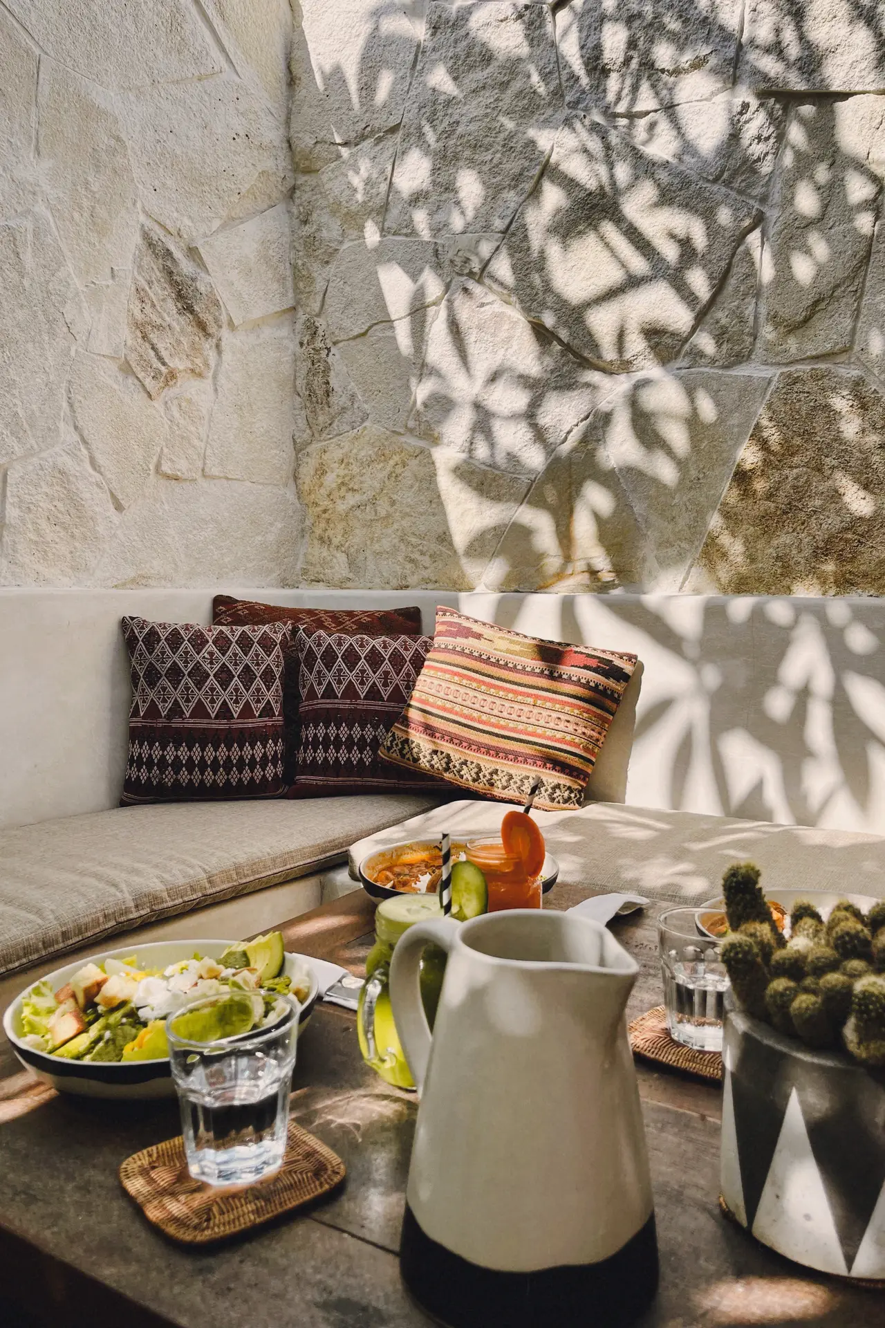 Cozy and sunlit corner seating area with dining table, perfect for relaxed outdoor dining. The space features textured stone walls, warm beige cushions, patterned throw pillows, and a rustic table set with fresh salads, drinks, and a ceramic pitcher. A stylish mix of natural tones and greenery makes this setup a great source of patio decor ideas for creating an inviting atmosphere.