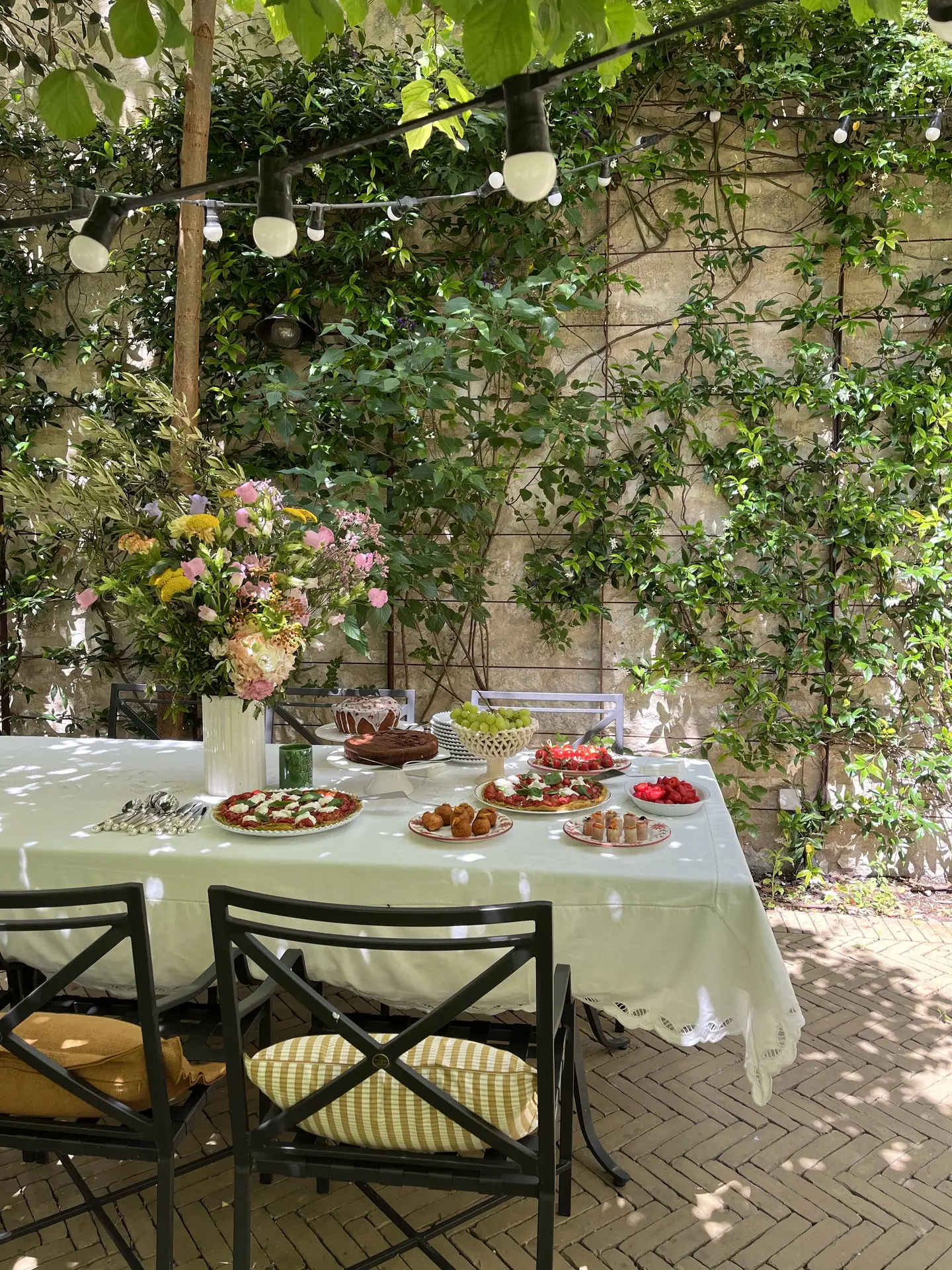 A beautiful outdoor setup featuring a dining table with greenery and fairy lights, surrounded by lush vines climbing up a stone wall. The table is elegantly dressed with a white tablecloth, floral centerpiece, and plates of colorful food - a perfect inspiration for patio decor ideas that blend nature and charm.