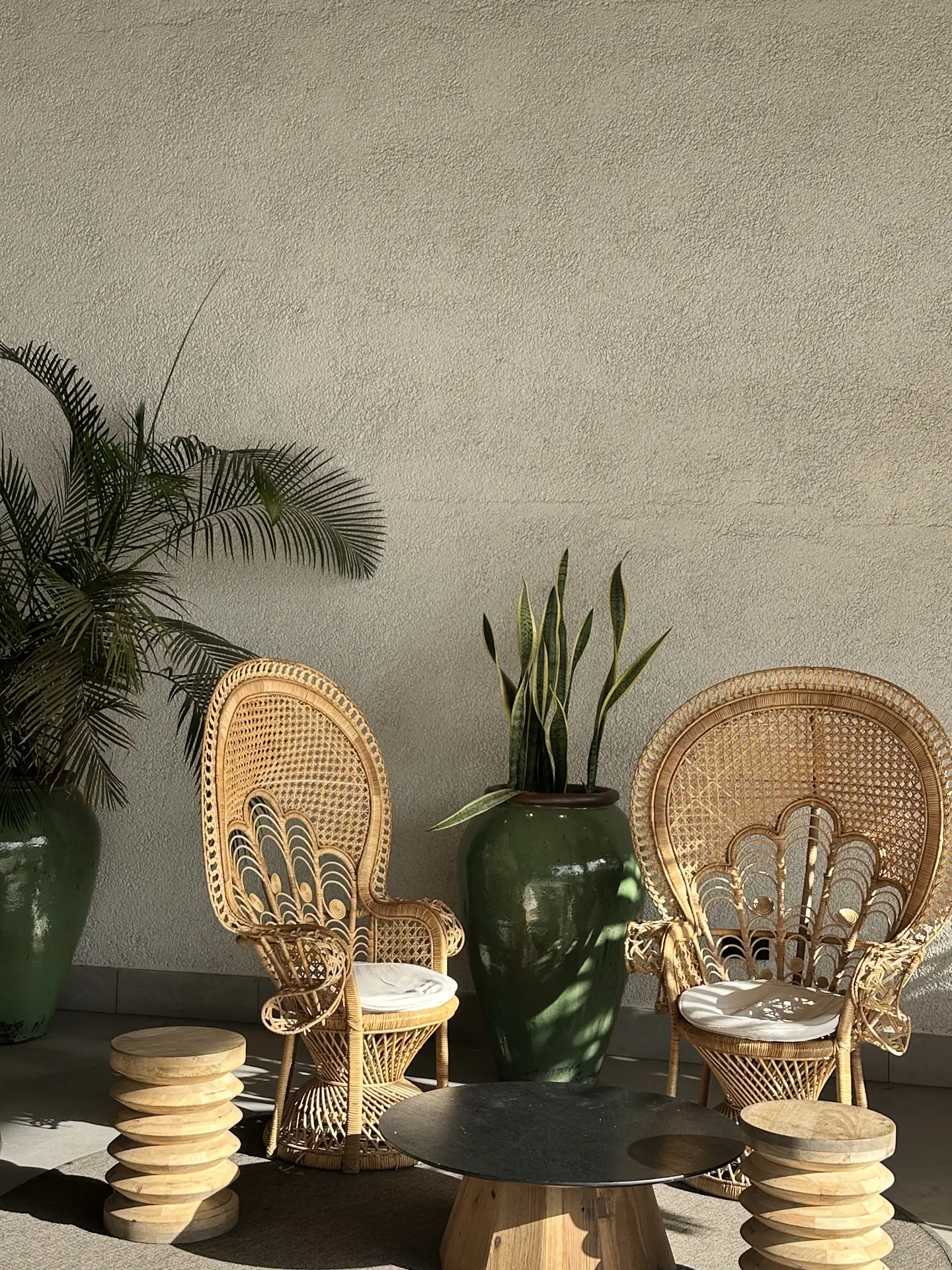 Stylish outdoor corner featuring extravagant wicker chairs with high curved backs, paired with a small round wooden table and sculptural stools. Lush green plants in oversized ceramic pots add life to the minimalist wall, offering elegant patio decor ideas that blend natural textures with sophisticated design.