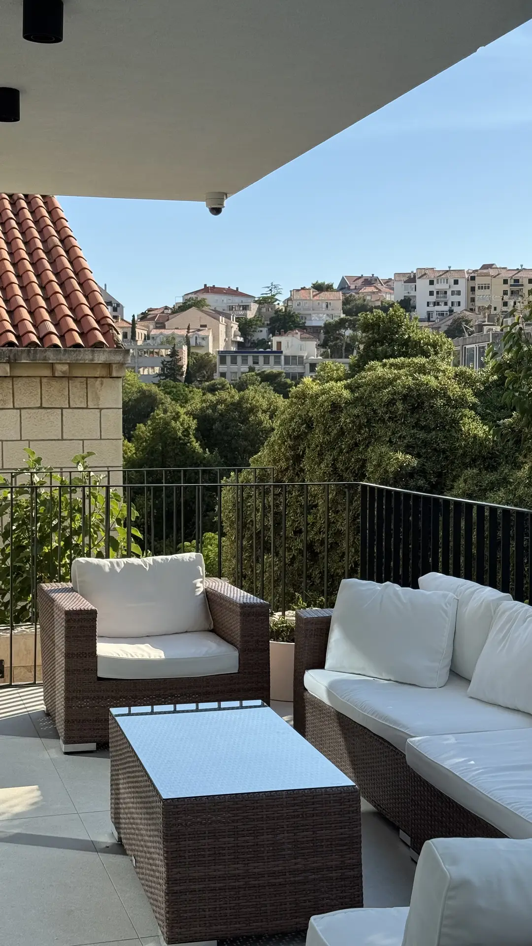 A stylish outdoor setup featuring patio decor ideas with an L-sectional and armchair made of wicker and topped with soft white cushions. The seating arrangement surrounds a sleek rectangular coffee table, set against a scenic backdrop of green trees and hillside homes under a clear blue sky.