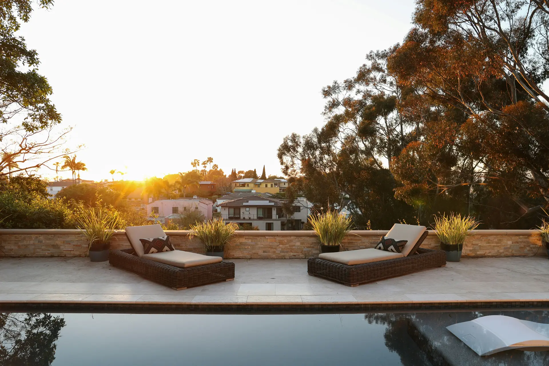 A serene outdoor setting showcasing patio decor ideas with two stylish woven loungers placed beside a tranquil pool. The scene is framed by potted greenery and warm sunset light, overlooking a hillside neighborhood with pastel-colored houses and tall trees glowing in the evening sun.