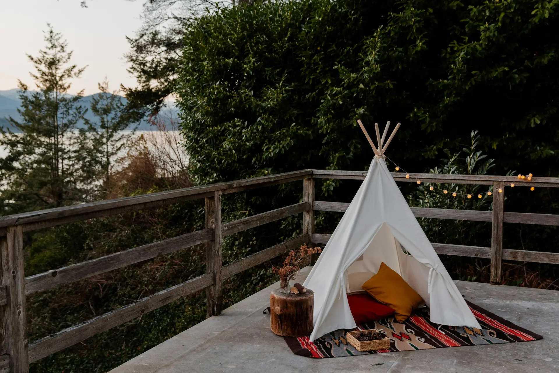 A cozy outdoor nook showcasing patio decor ideas with a charming play tent for the kids set on a colorful patterned rug. Soft pillows in mustard and beige add comfort, while string lights and a small wooden stump table with a vase bring warmth and character to this nature-surrounded patio overlooking a scenic view.
