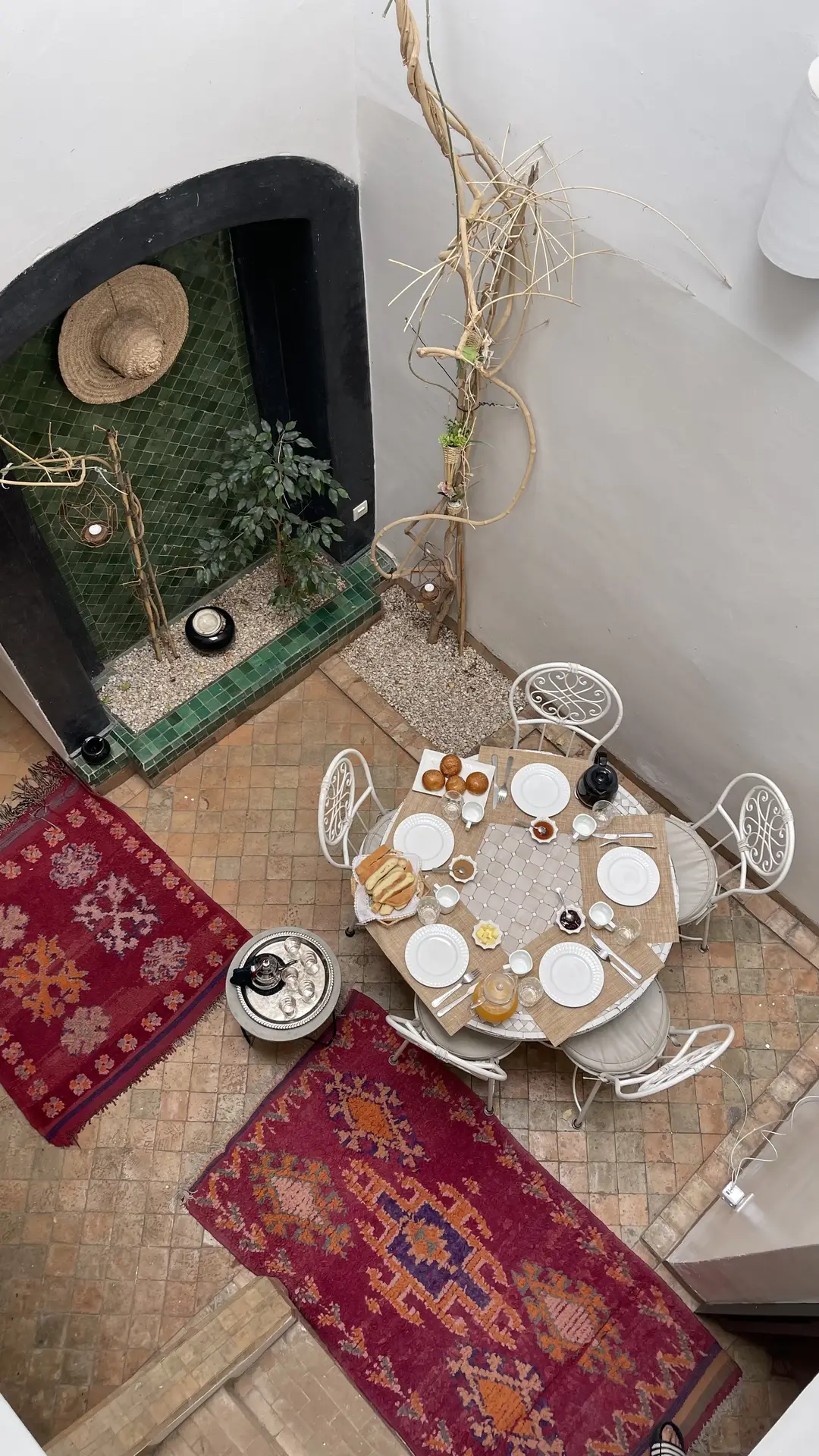 Cozy courtyard with a round dining table set for breakfast, surrounded by white wrought-iron chairs and colorful patterned rugs for boho vibes. The space features earthy tiles, a green-tiled wall with plants, and natural decor elements, a charming setup full of patio decor ideas that blend rustic elegance with relaxed comfort.