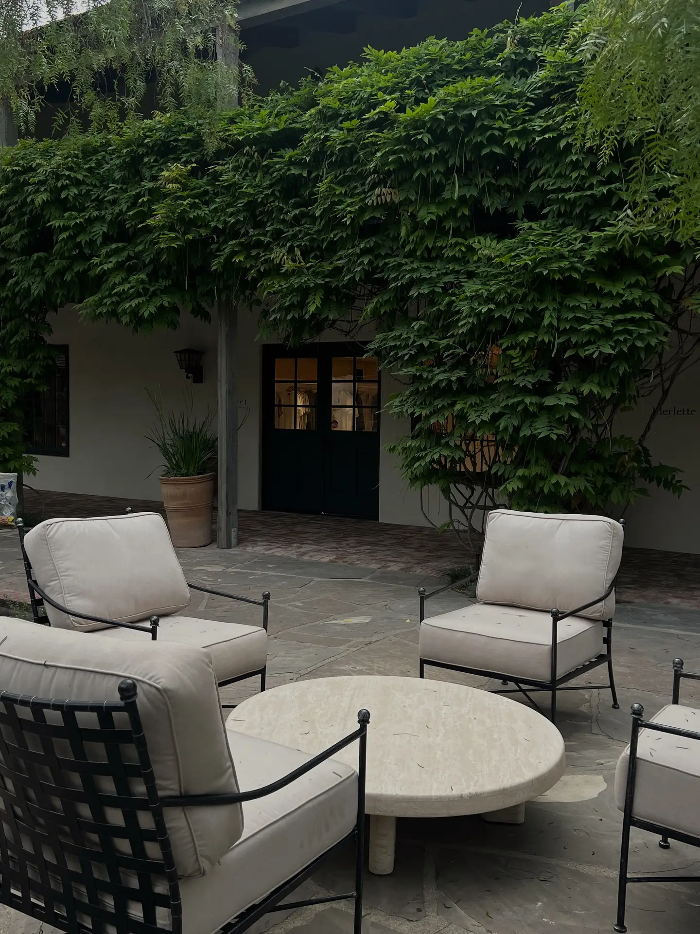 A cozy courtyard setting featuring patio decor ideas with several armchairs around a low table. The cream-cushioned chairs with black metal frames form a relaxed conversation area on a stone patio, surrounded by lush green climbing plants that cover the walls and frame the dark wooden doors in the background.