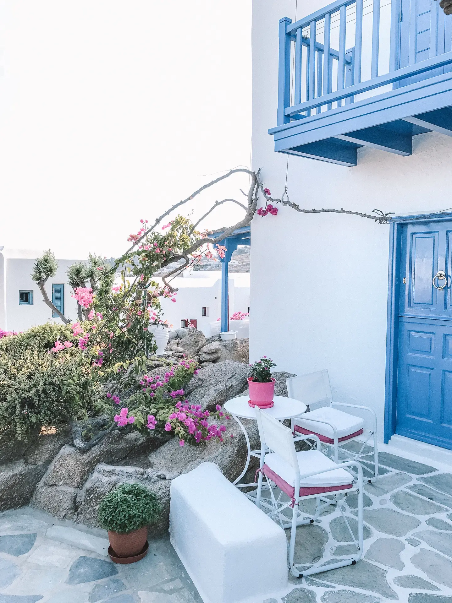 A bright Mediterranean-inspired patio showcasing patio decor ideas with a small seating area of two white chairs and a round table beside a blue door. Pink bougainvillea climbs along white stone walls, while blue balcony railings and shutters add a fresh coastal touch under the sunny sky.