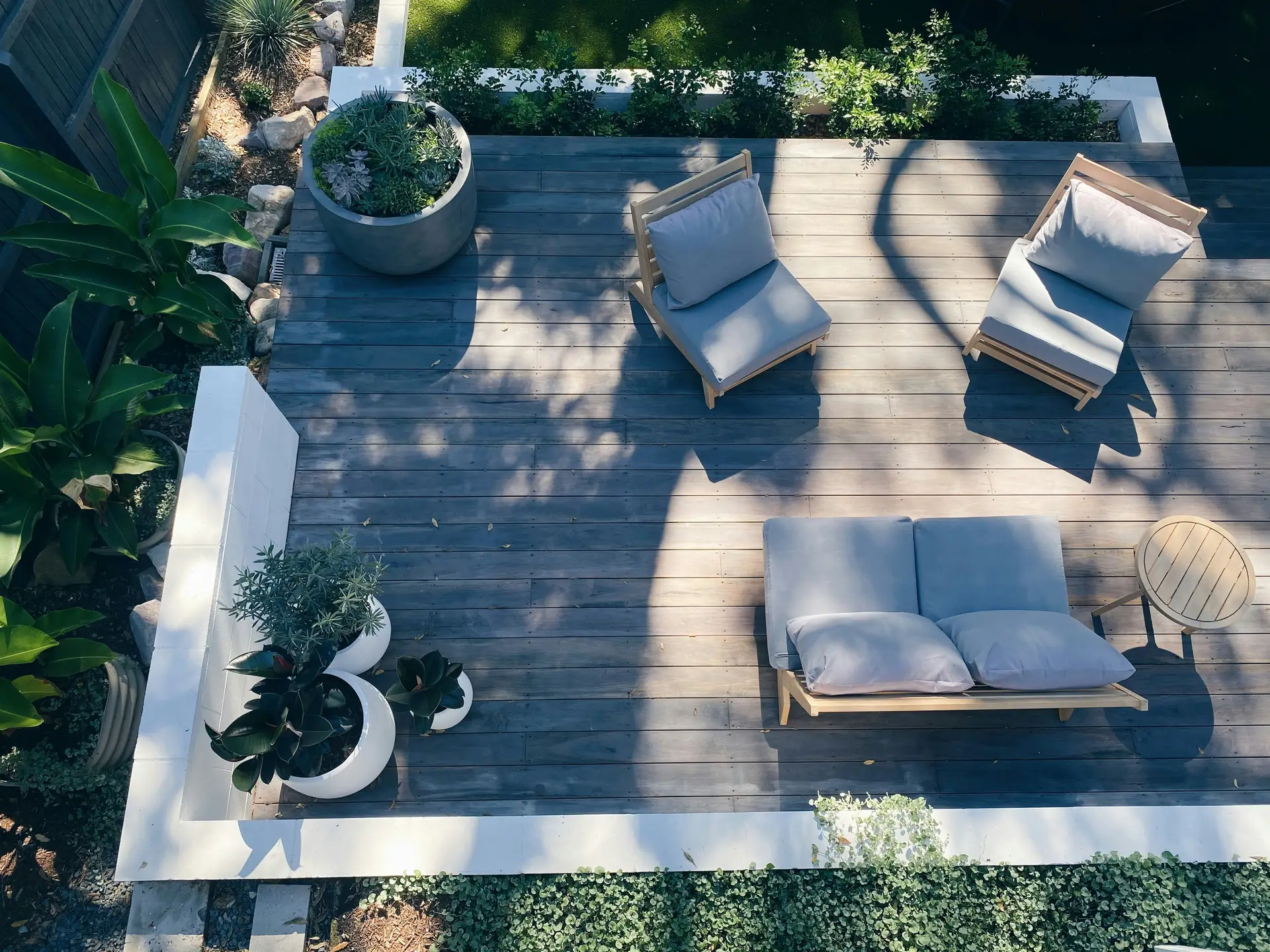 A serene outdoor space showcasing patio decor ideas with a small sofa and two armchairs arranged on a wooden deck. The neutral-toned seating blends seamlessly with the natural surroundings, complemented by modern planters filled with lush greenery. The design creates a calm, open-air retreat ideal for lounging or casual conversation.