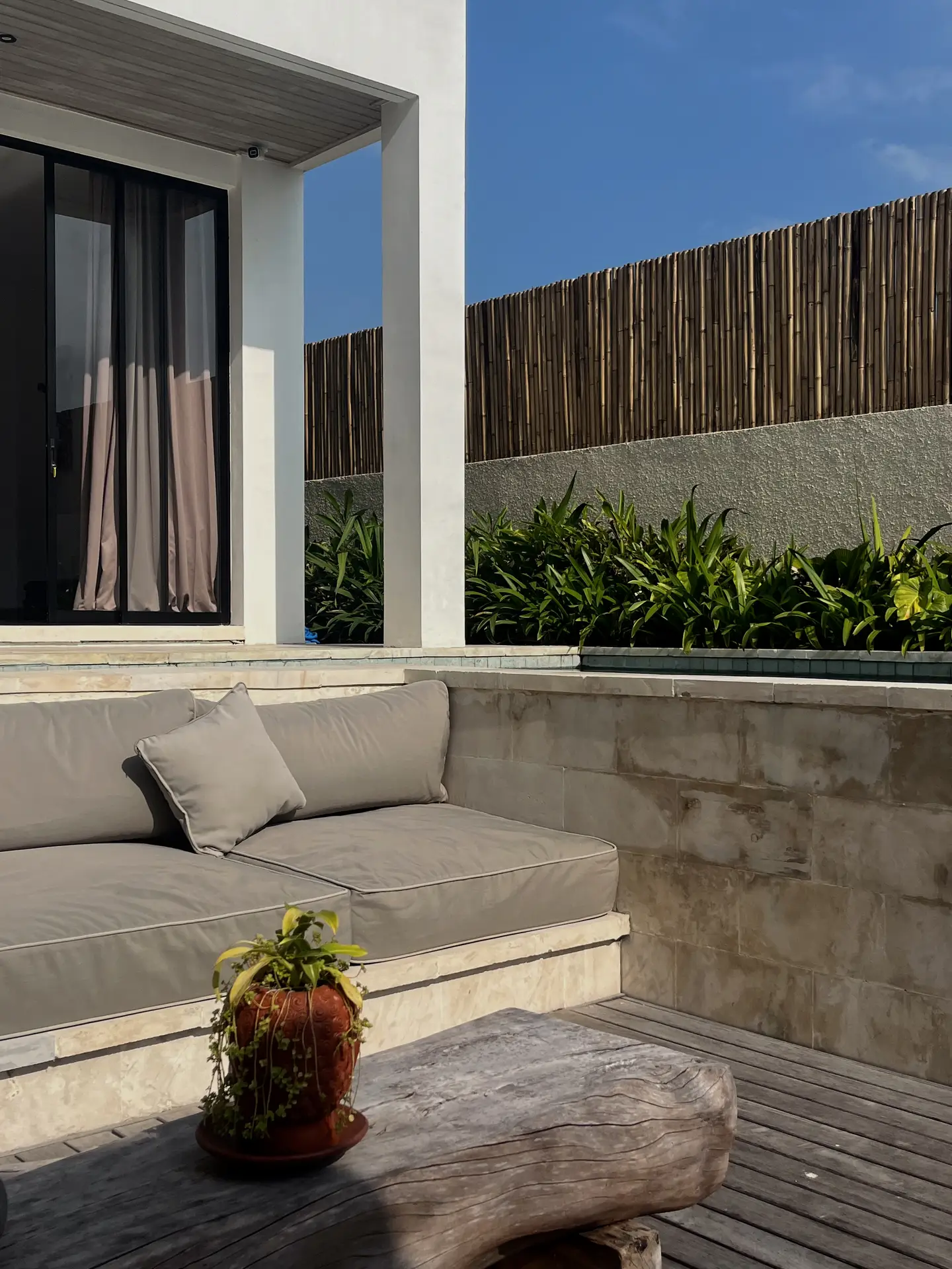 A minimalist outdoor lounge showcasing patio decor ideas with a sofa integrated into stone with wooden bench as table. The space features neutral-toned cushions, a rustic wooden centerpiece, and lush greenery against a bamboo privacy fence, creating a serene and stylish retreat under a clear blue sky.