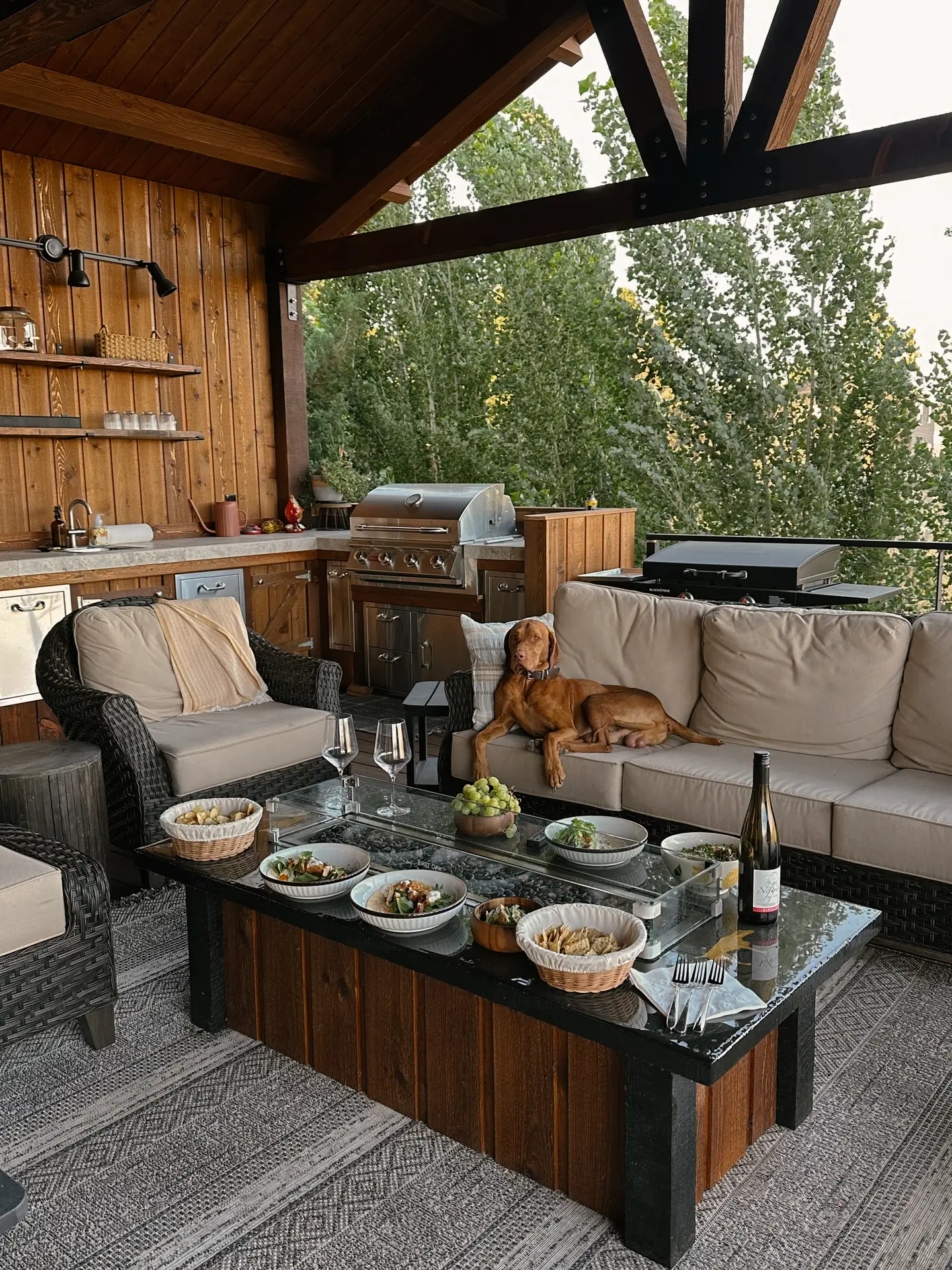 A cozy outdoor setup featuring patio decor ideas with a sofa with arm chairs and a low table under a wooden pergola. The scene includes a dog lounging on the couch, a table set with dinner bowls and wine glasses, and a built-in grill area in the background-perfect for relaxed evenings surrounded by nature.