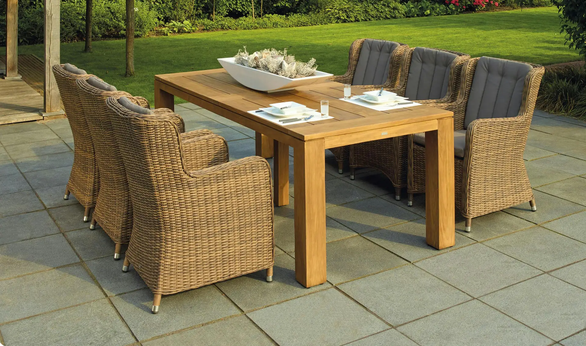 A stylish outdoor setup showcasing patio decor ideas with a sturdy wooden dining table with symmetrically arranged chairs made of wicker and soft gray cushions. The clean lines, balanced layout, and natural materials create a cozy yet elegant atmosphere against a lush green garden backdrop.