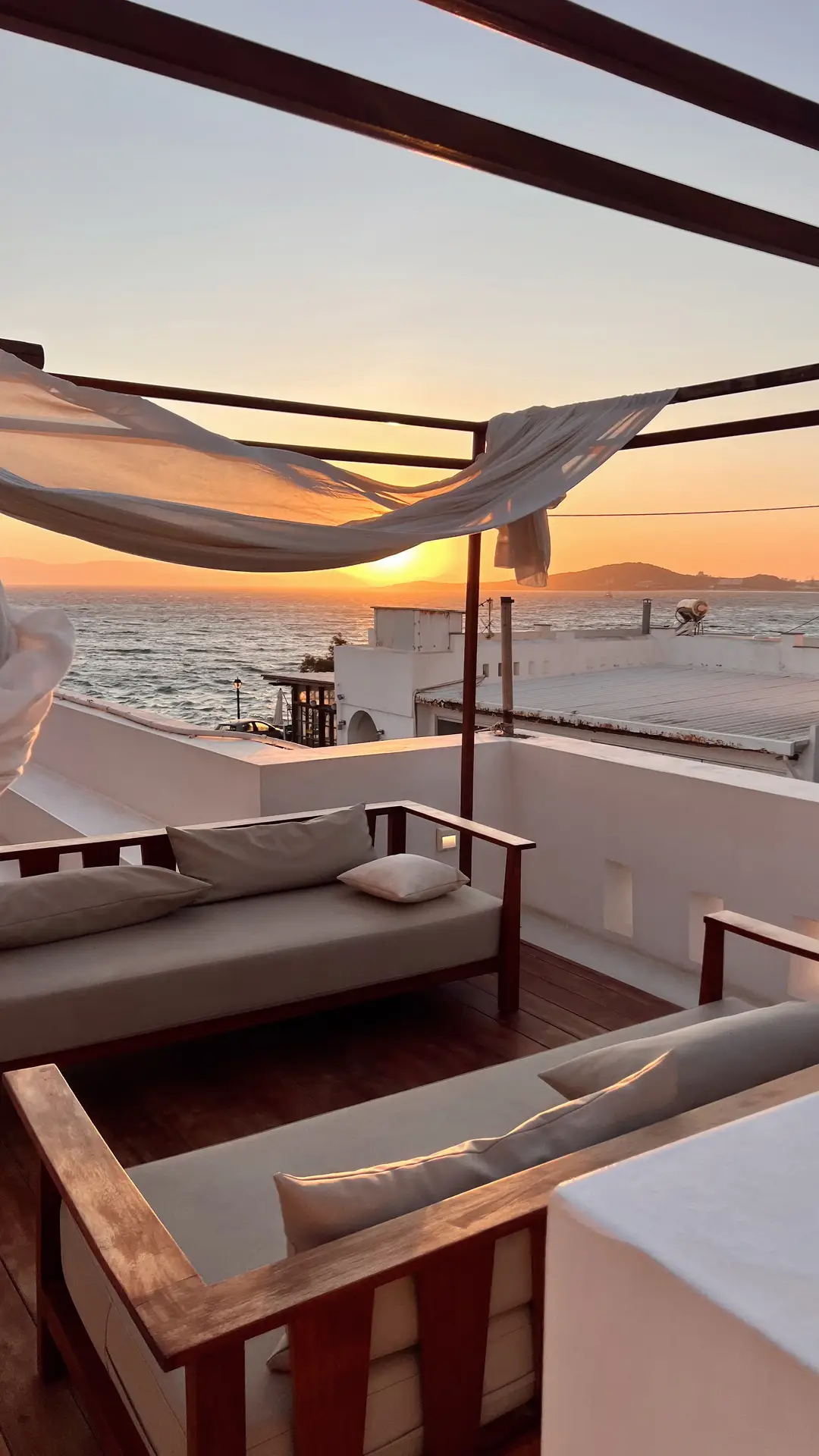 Serene coastal terrace at sunset with two sofas facing each other under a wooden pergola draped in flowing white fabric. The soft golden light reflects off the ocean, enhancing the warm wood tones and minimalist cushions. A stunning scene that captures effortless elegance and relaxation, perfect for patio decor ideas that combine comfort with a view.