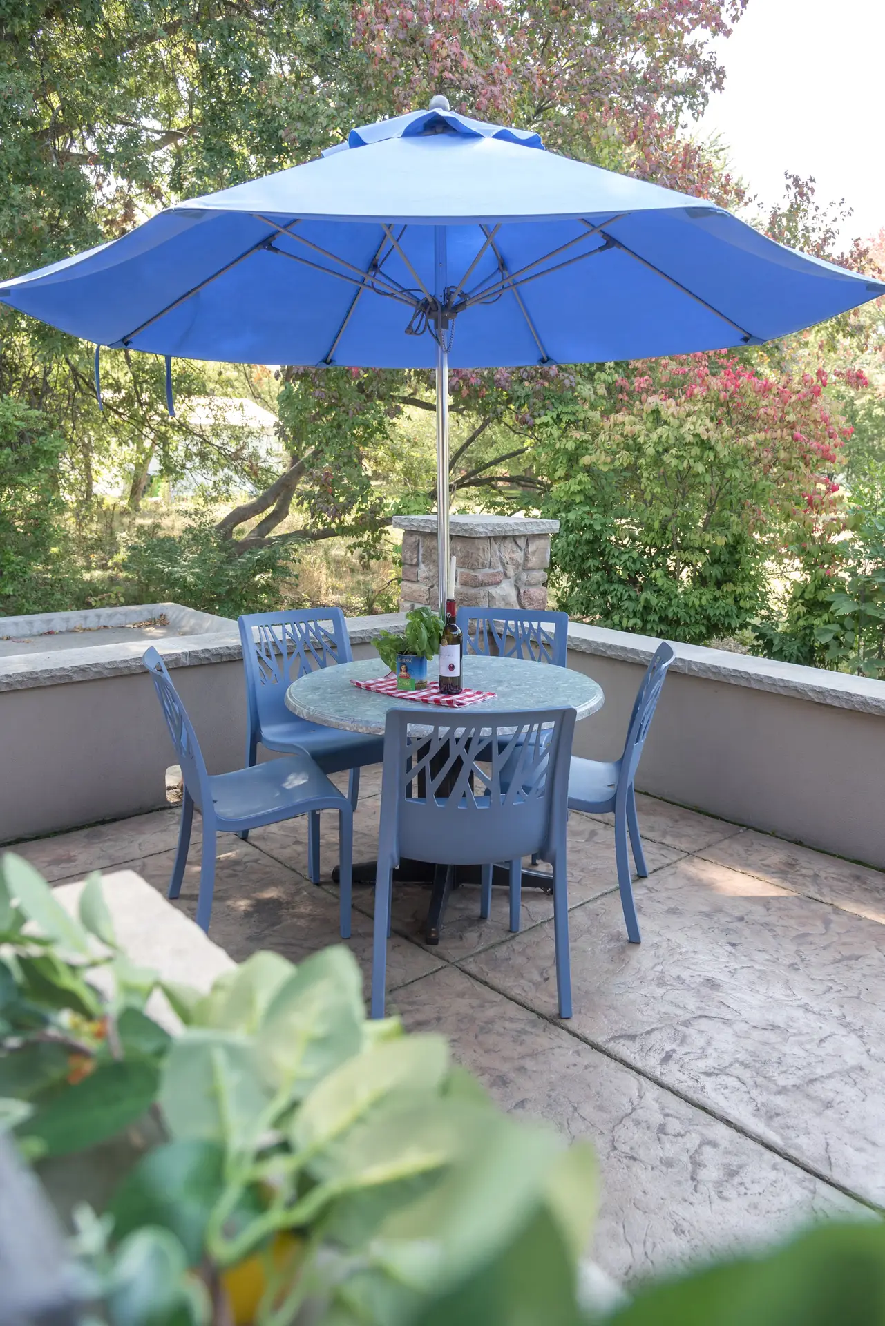 A stylish outdoor setup showcasing patio decor ideas with uniform colors, blue chairs surrounding a matching round table under a large blue umbrella. The cohesive color palette creates a calm and inviting atmosphere, perfect for relaxed outdoor dining amid lush green trees and a peaceful garden view.