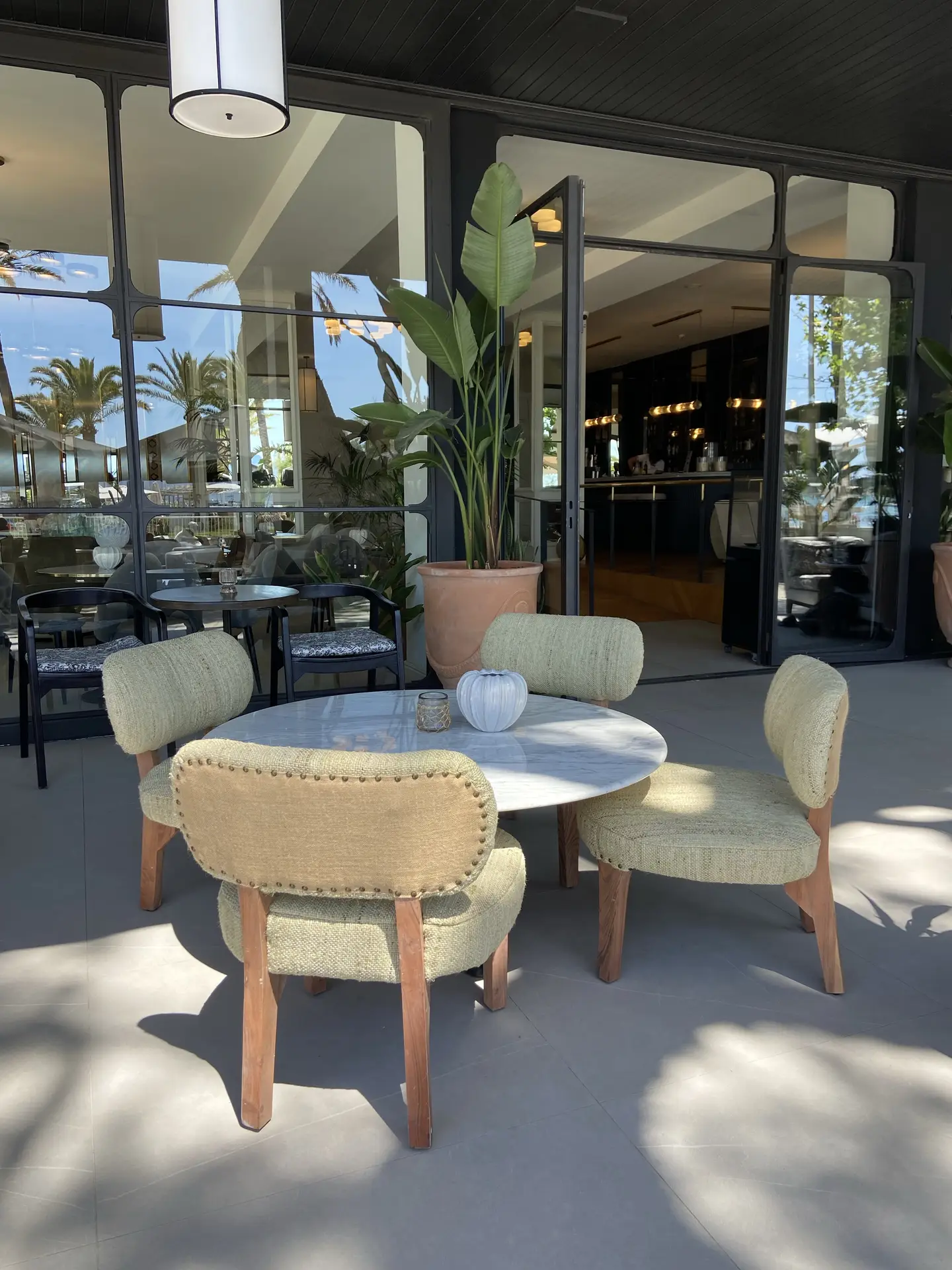 Stylish outdoor terrace with upholstered chairs around a round table, framed by tall potted plants and large glass doors leading inside. The neutral tones, soft sunlight, and lush greenery create an elegant and relaxing atmosphere, perfect for anyone seeking modern patio decor ideas that balance comfort and sophistication.
