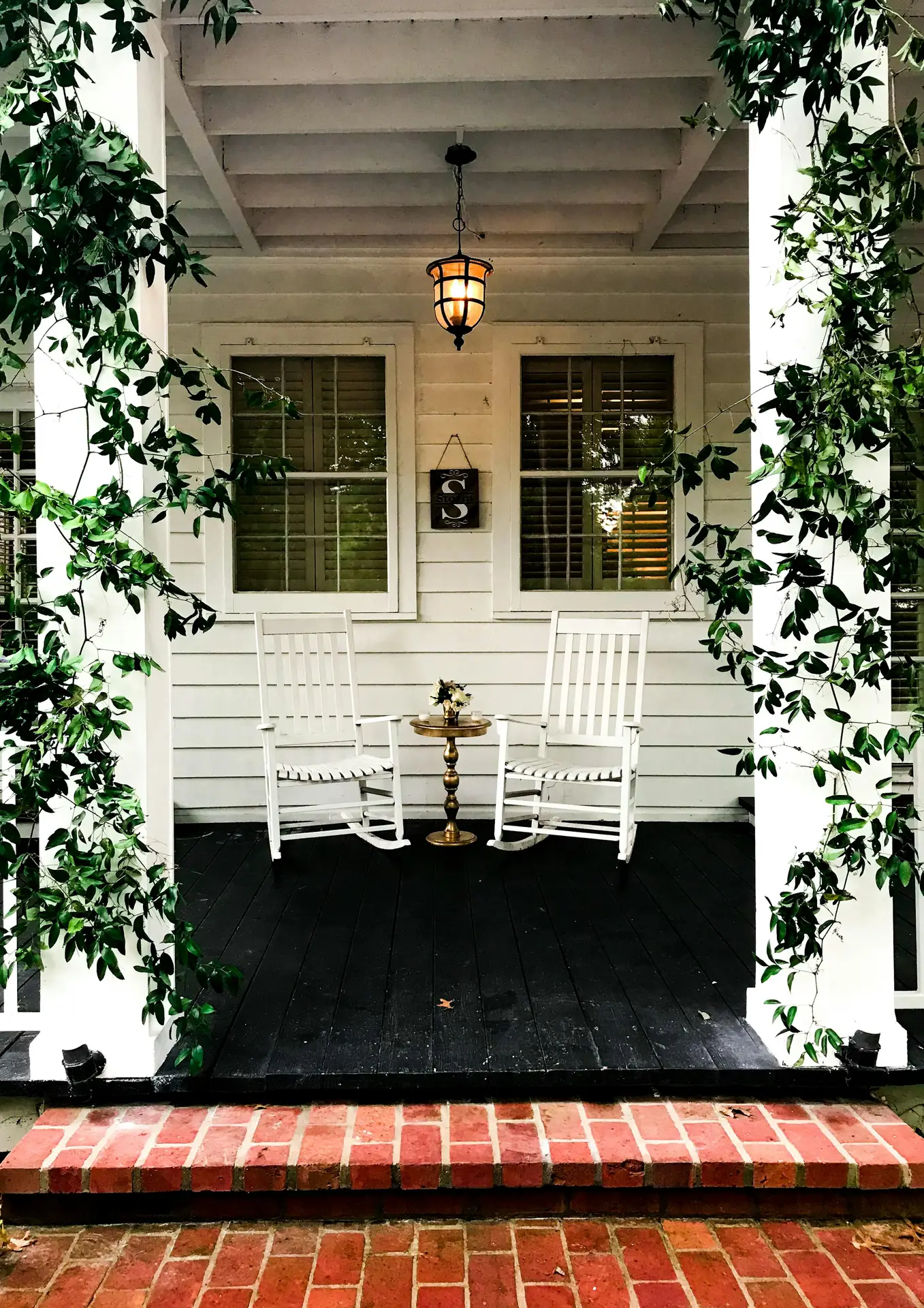 A charming front porch setup featuring white rocking chairs and a small brass side table, surrounded by climbing greenery and soft evening light. The mix of brick steps, black flooring, and white paneling offers timeless patio decor ideas that feel cozy and welcoming.